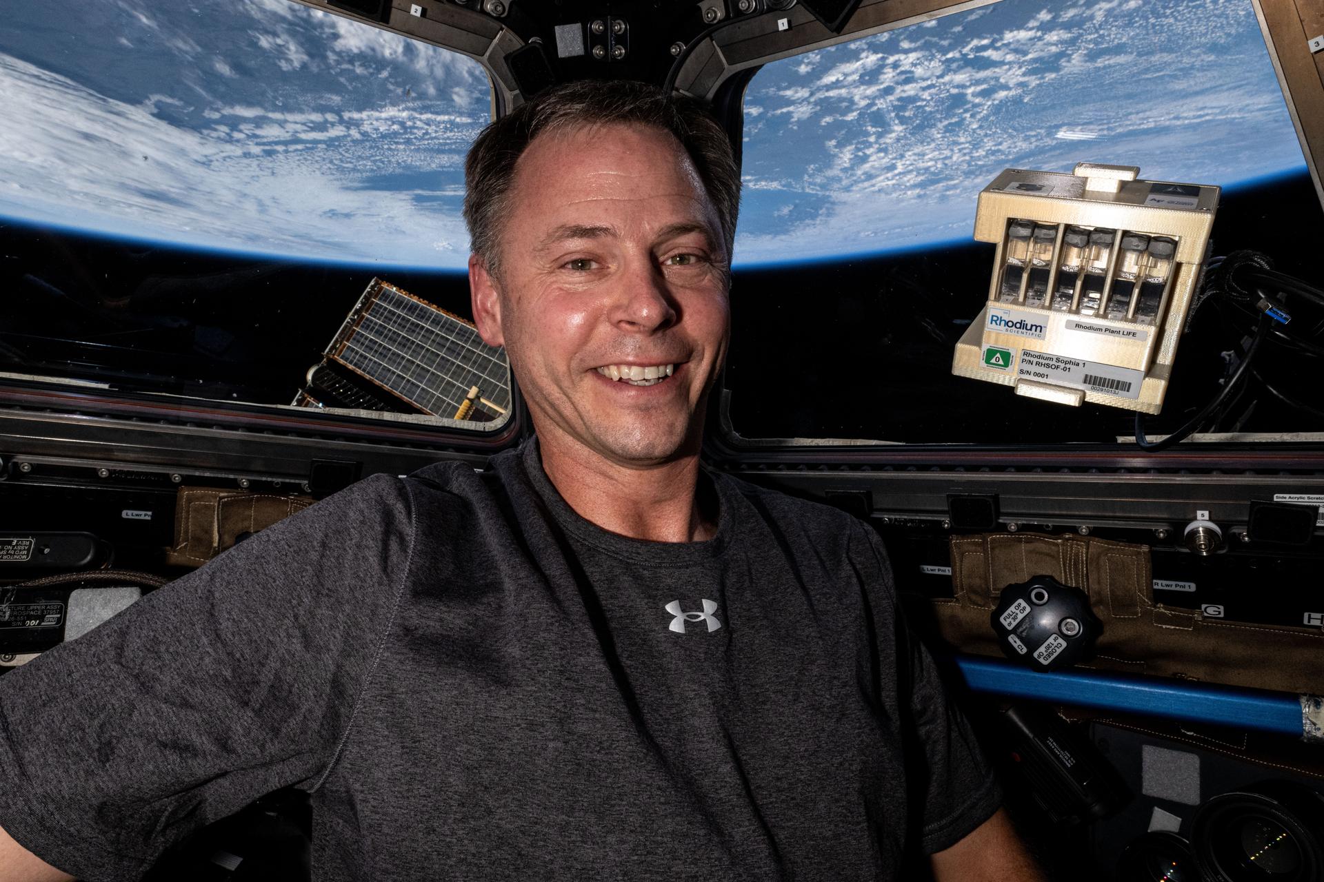 Hague, wearing a gray t-shirt and smiling at the camera, is inside the cupola with hardware for the Rhodium Plant LIFE investigation. To his right floats the hardware, a shiny gold box that holds six glass sample tubes with a white sticker that says “Rhodium.” Earth is visible through the cupola windows in the background.
