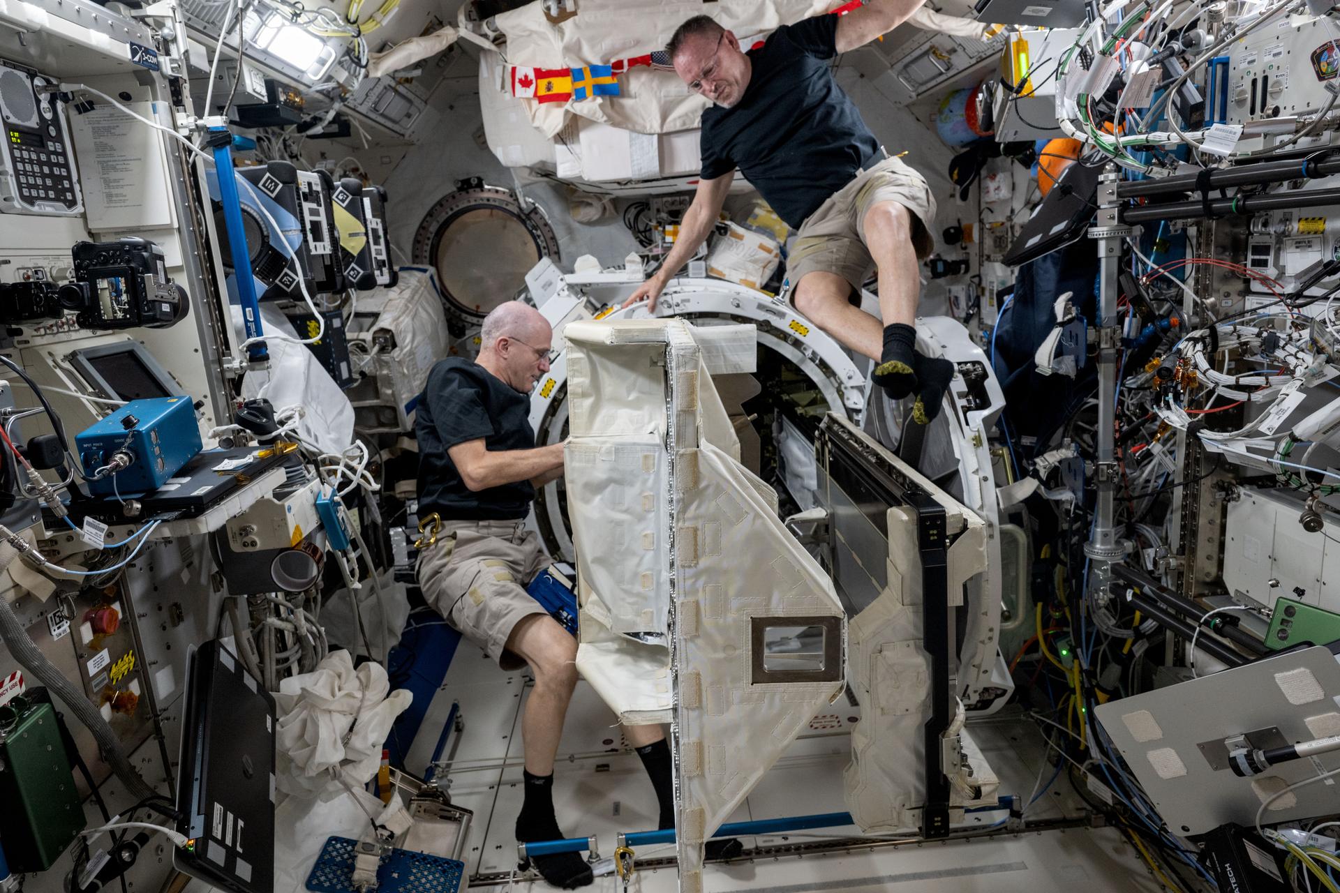 Expedition 72 Flight Engineers (from left) Don Pettit and Butch Wilmore, both NASA astronauts, remove a small satellite deployer from inside the Kibo laboratory module's airlock that had earlier deployed several CubeSats into Earth orbit for a series of technology studies.