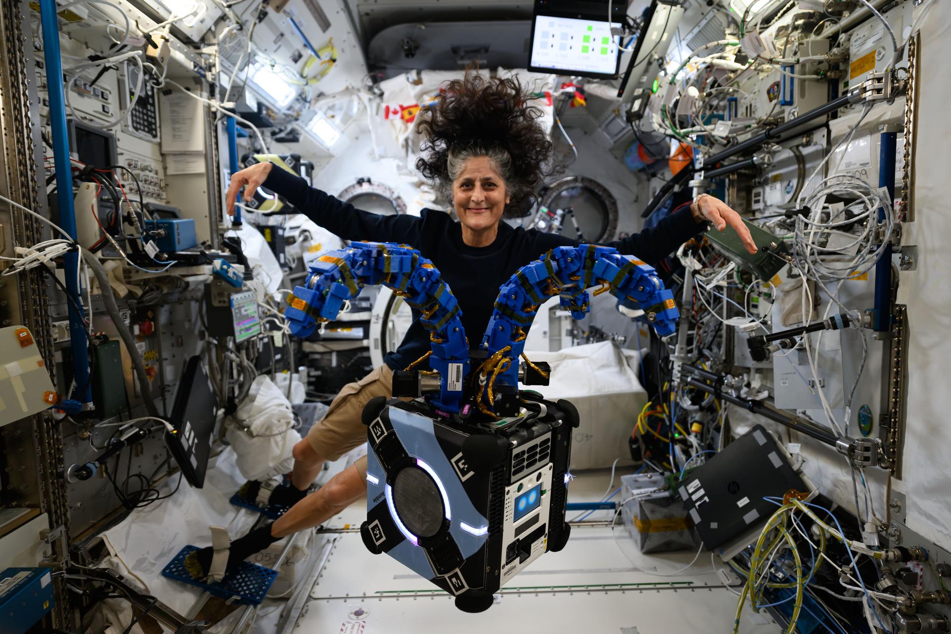 NASA Astronaut Suni Williams floats inside the International Space Station with her hair extending outward due to microgravity. She is interacting with the Astrobee robotic assistant featuring two articulated blue robotic arms attached to a cube-shaped device. The background shows an intricate array of scientific equipment, wiring, and monitors, capturing the high-tech environment of the space station.