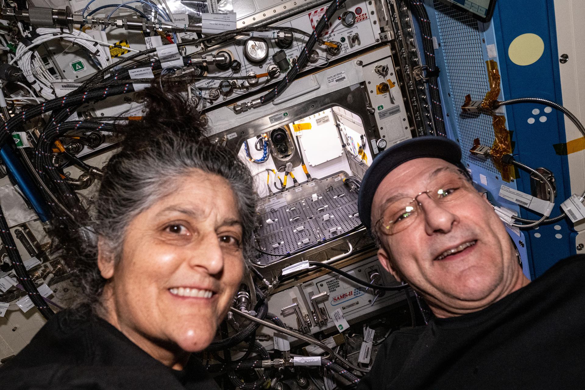 Expedition 72 Commander Suni Williams and Flight Engineer Don Pettit, both NASA astronauts, pose for a portrait in front of the Advanced Plant Habitat located inside the International Space Station's Kibo laboratory module. The Plant Habitat provides a large, enclosed, environmentally controlled chamber designed to support commercial and fundamental plant research in microgravity.