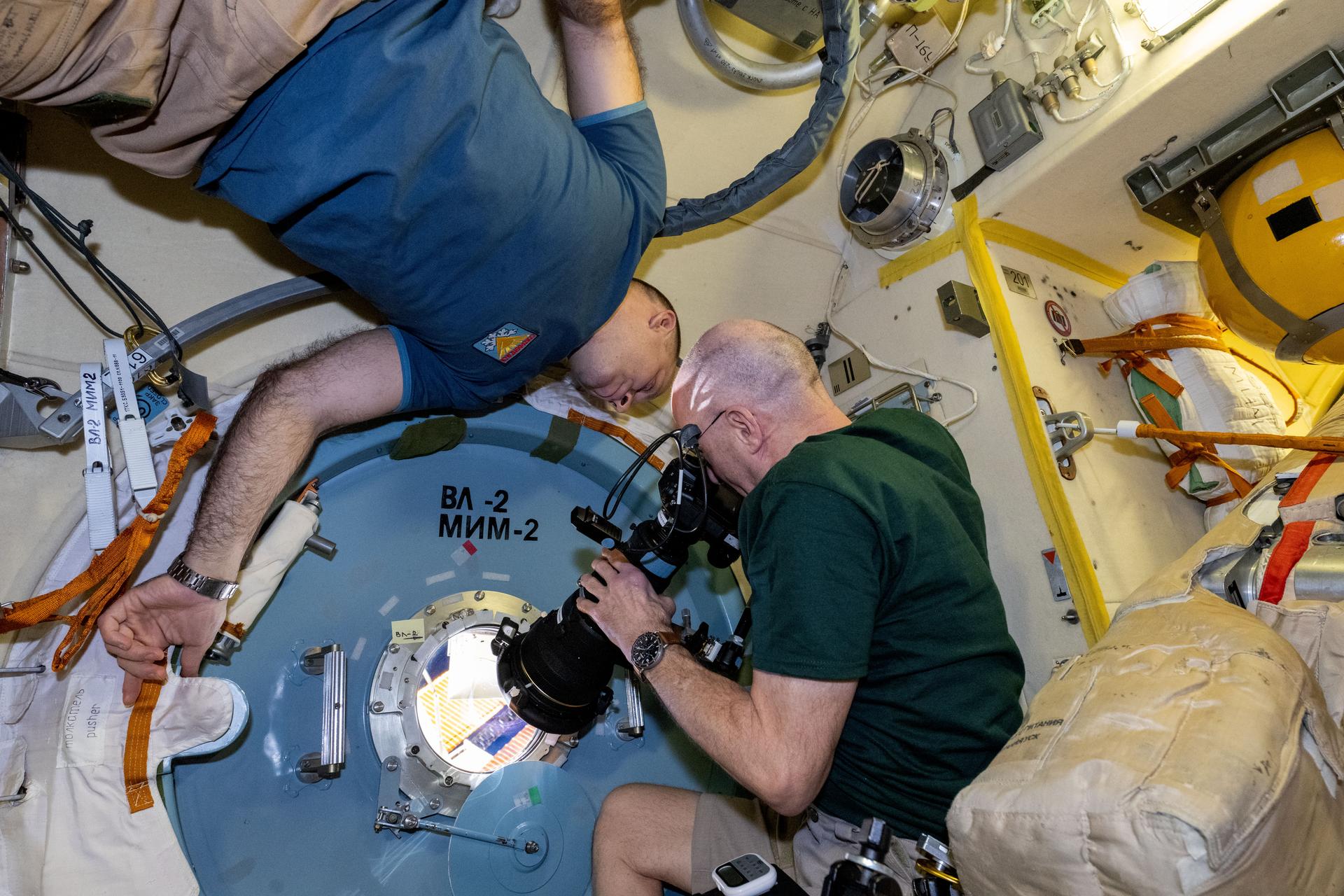 NASA astronaut and Expedition 72 Flight Engineer Don Pettit points a camera outside a window on the International Space Station's Poisk module for a sun photography session with assistance from Roscosmos cosmonaut and Expedition 72 Flight Engineer Ivan Vagner.