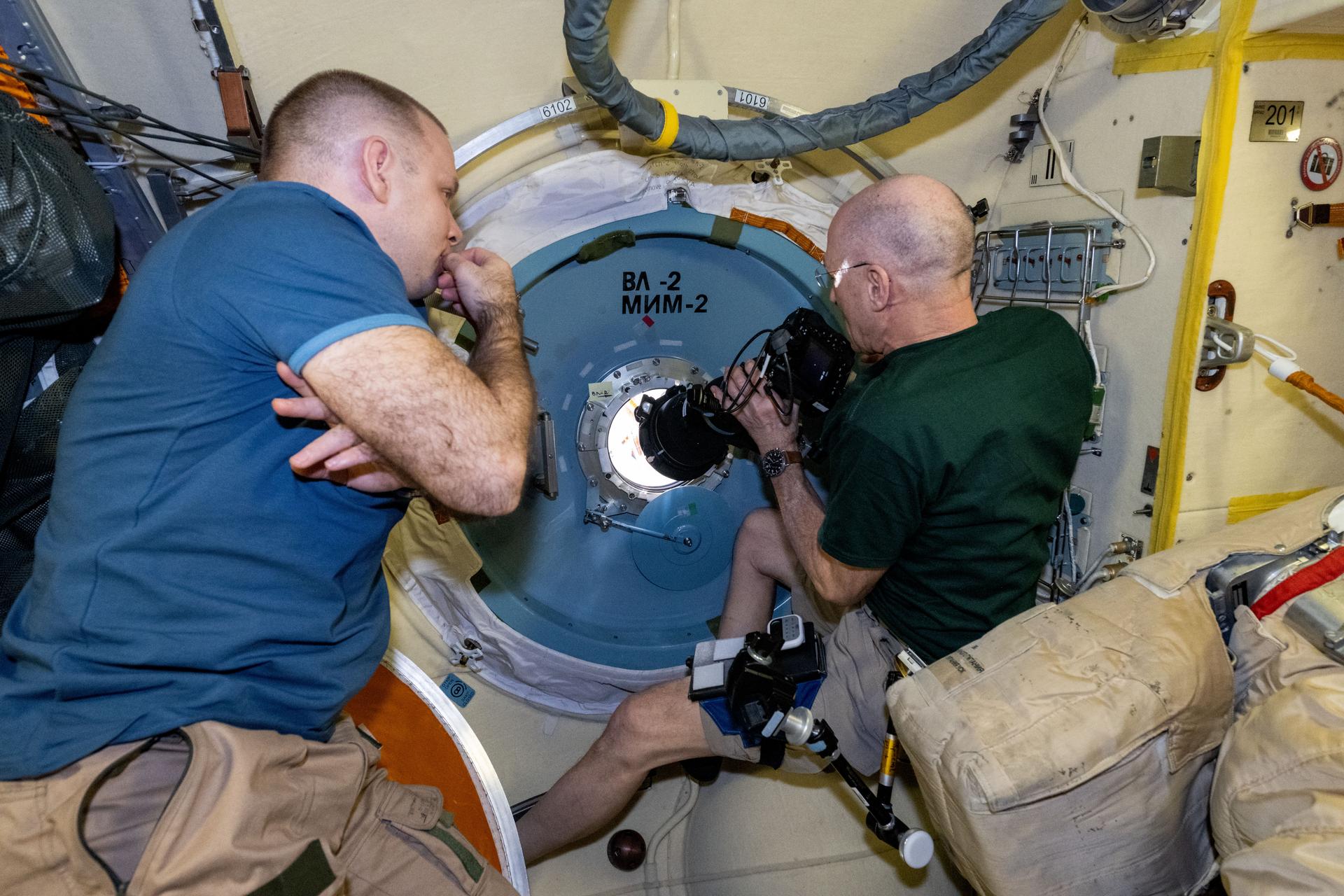 NASA astronaut and Expedition 72 Flight Engineer Don Pettit points a camera outside a window on the International Space Station's Poisk module for a sun photography session with assistance from Roscosmos cosmonaut and Expedition 72 Flight Engineer Ivan Vagner.