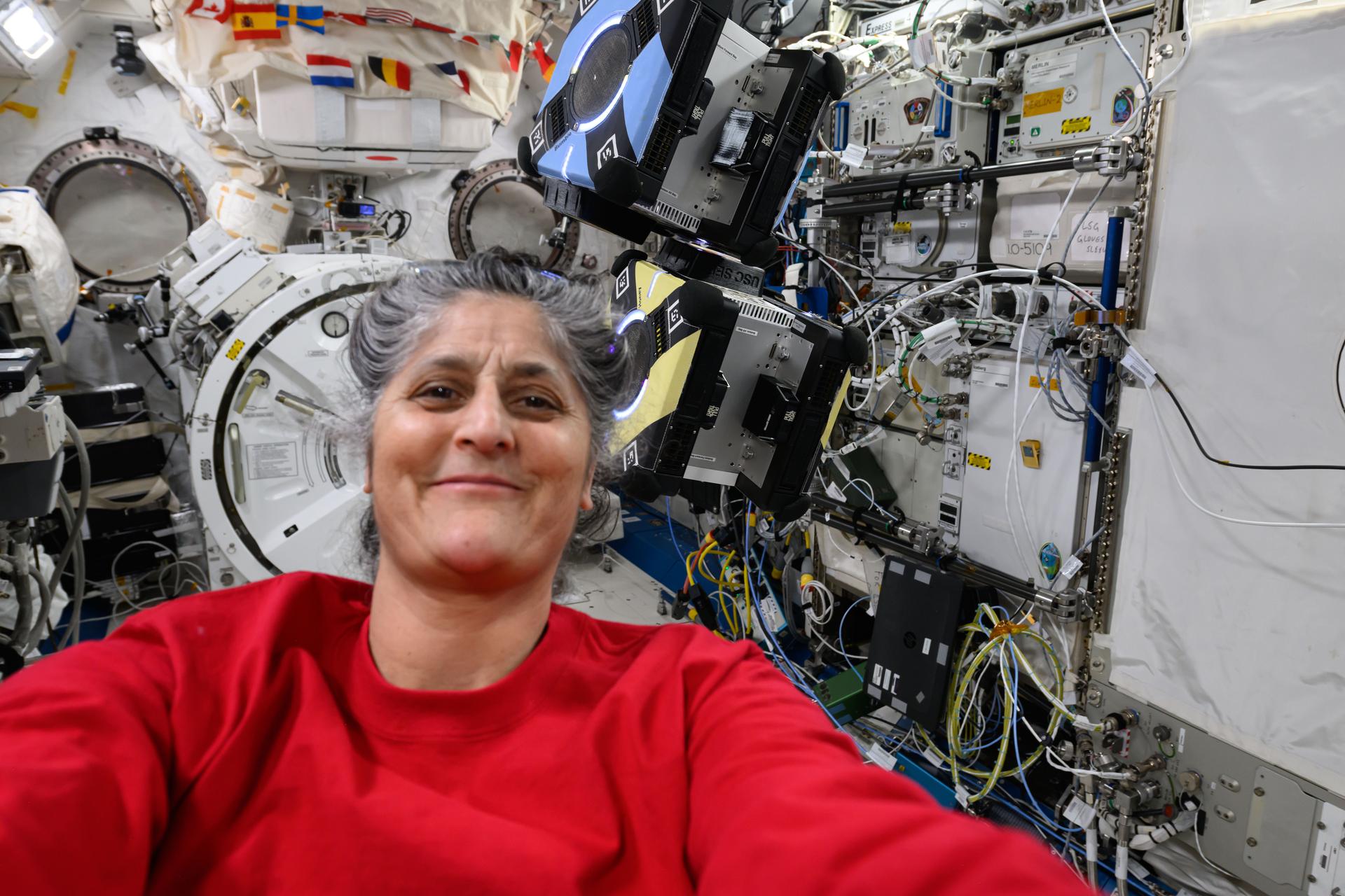 NASA astronaut and Expedition 72 Commander Suni Williams takes a selfie portrait with a pair of Astrobee robotic free-flyers behind her inside the International Space Station's Kibo laboratory module.
