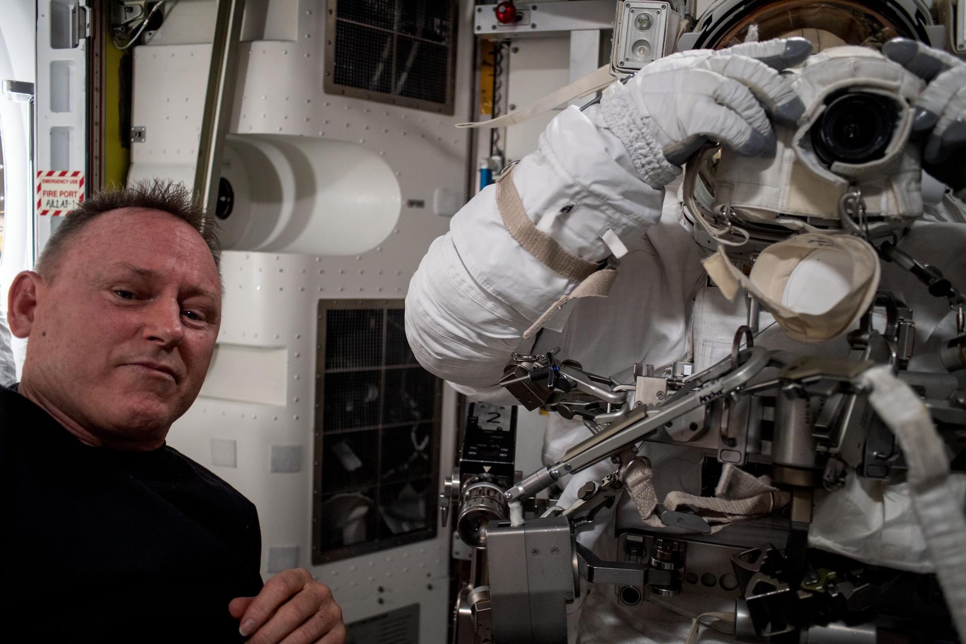 NASA astronaut and Expedition 72 Flight Engineer Butch Wilmore (left) assists NASA astronaut Nick Hague (right) as he tries on and evaluates his spacesuit in a pressurized configuration aboard the International Space Station's Quest airlock. Hague is pictured holding a spacewalking camera in front of his spacesuit's helmet shield.