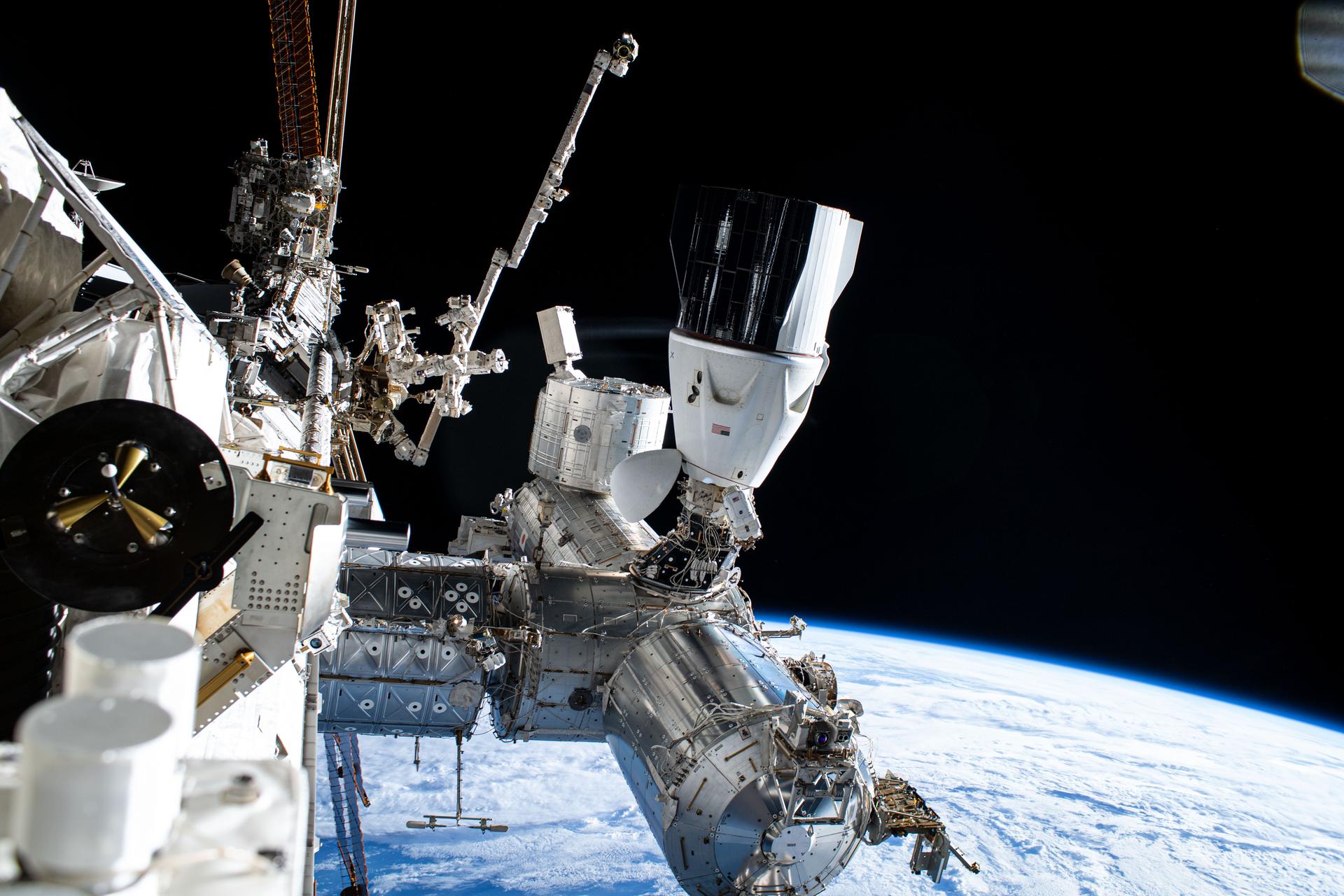 NASA astronaut and Expedition 72 Commander Suni Williams (center) is dwarfed near the SpaceX Dragon crew spacecraft as she replaces a planar reflector, advanced navigational hardware visiting vehicles use when approaching the International Space Station. Dragon is docked to the Harmony module's space-facing port which rests in between the Kibo and Columbus laboratory modules. 267 miles below is the Pacific Ocean east of New Zealand.