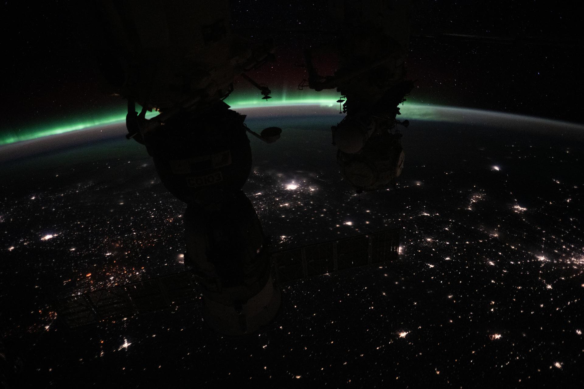 The aurora borealis crowns Earth's horizon with the city lights of Canada dotting the landscape below in this photograph from the International Space Station as it orbited 262 miles above North America. (From left) The Soyuz MS-26 spacecraft and the Pirs docking compartment silhouette the foreground.
