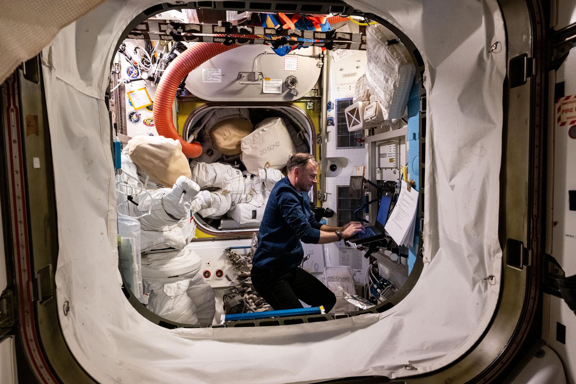 NASA astronaut and Expedition 72 Flight Engineer Nick Hague works on a laptop computer inside the International Space Station's Quest airlock with a pair of spacesuits staged behind him.
