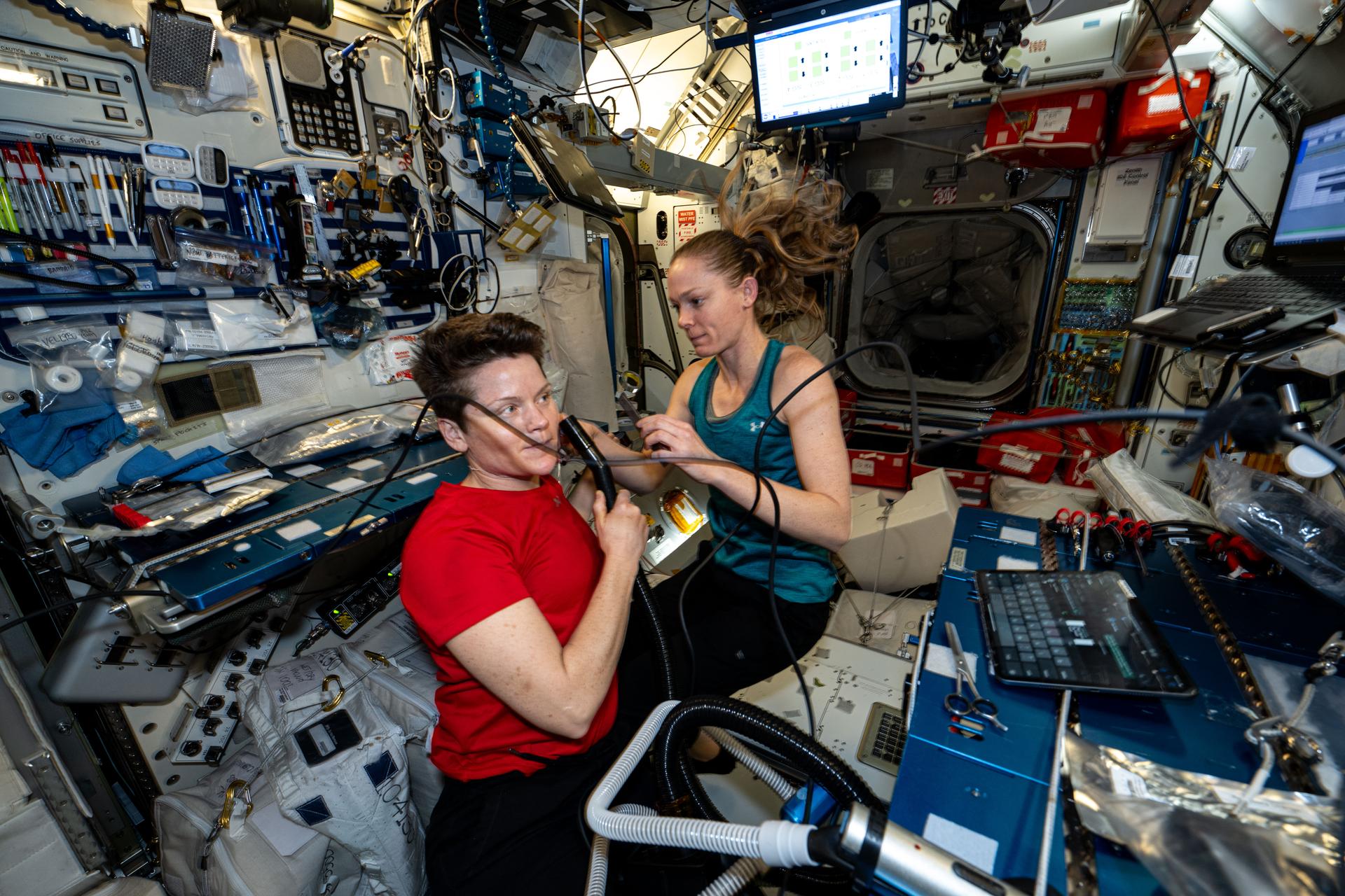 NASA astronauts (left to right) Anne McClain and Nichole Ayers, both Expedition 73 Flight Engineers, are pictured inside the International Space Station's Harmony module. Ayers trims McClain's hair as a suction hose collects loose hairs to protect the station's atmosphere.