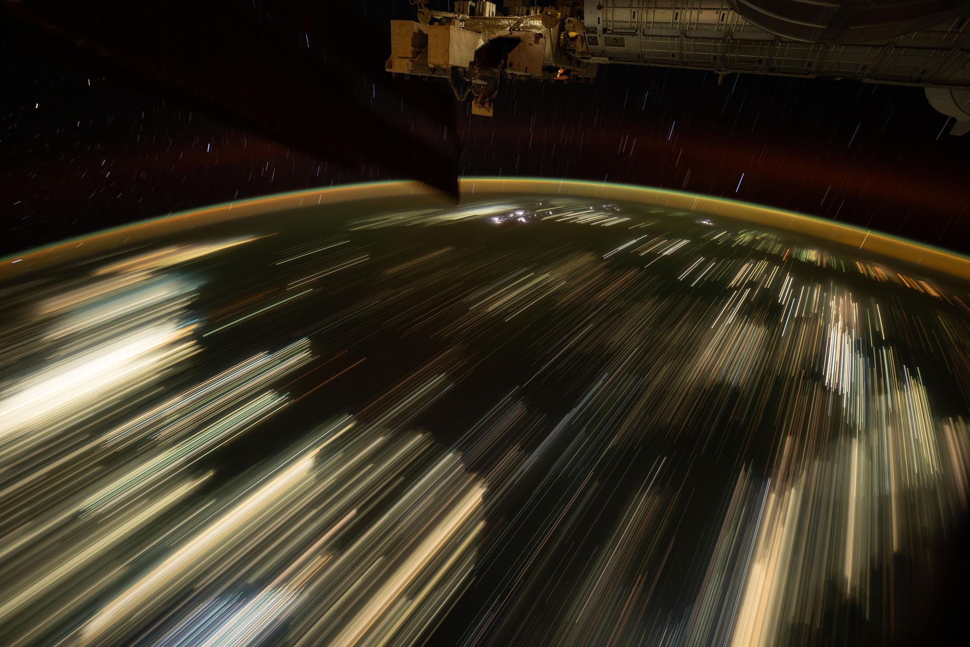 This long-duration photograph taken from the International Space Station orbiting 259 miles above the South China Sea highlights the city lights of Southeast Asia streaking below, a lightning storm near the top of Earth's horizon, and star trails above an atmopsheric glow.
