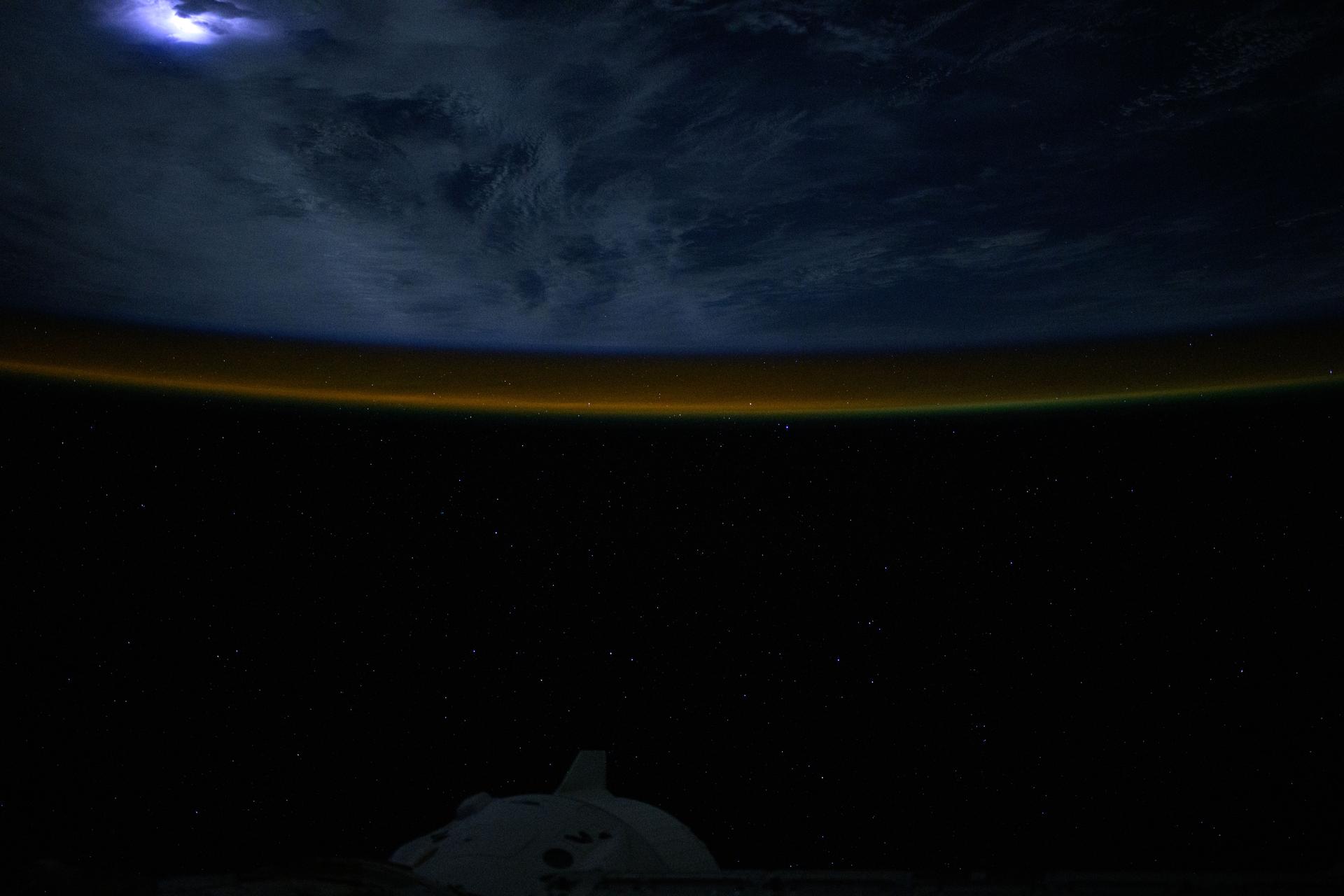 Lightning (upper left) illuminates the clouds and an atmospheric glow blankets Earth's horizon in this photograph from the International Space Station as it orbited 264 miles above the Atlantic Ocean off the coast of southern Brazil. At bottom, is the SpaceX Dragon crew spacecraft docked to the orbital outpost's forward port on the Harmony module.