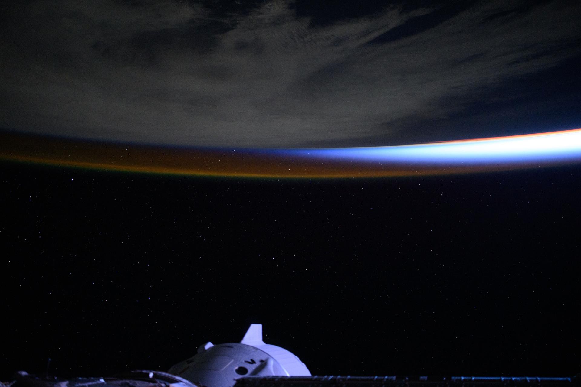 An orbital sunrise breaks above a cloudy Atlantic Ocean and intersects with Earth's atmospheric glow in this photograph from the International Space Station as it orbited 268 miles overhead. At bottom, is the SpaceX Dragon crew spacecraft docked to the orbital outpost's forward port on the Harmony module.