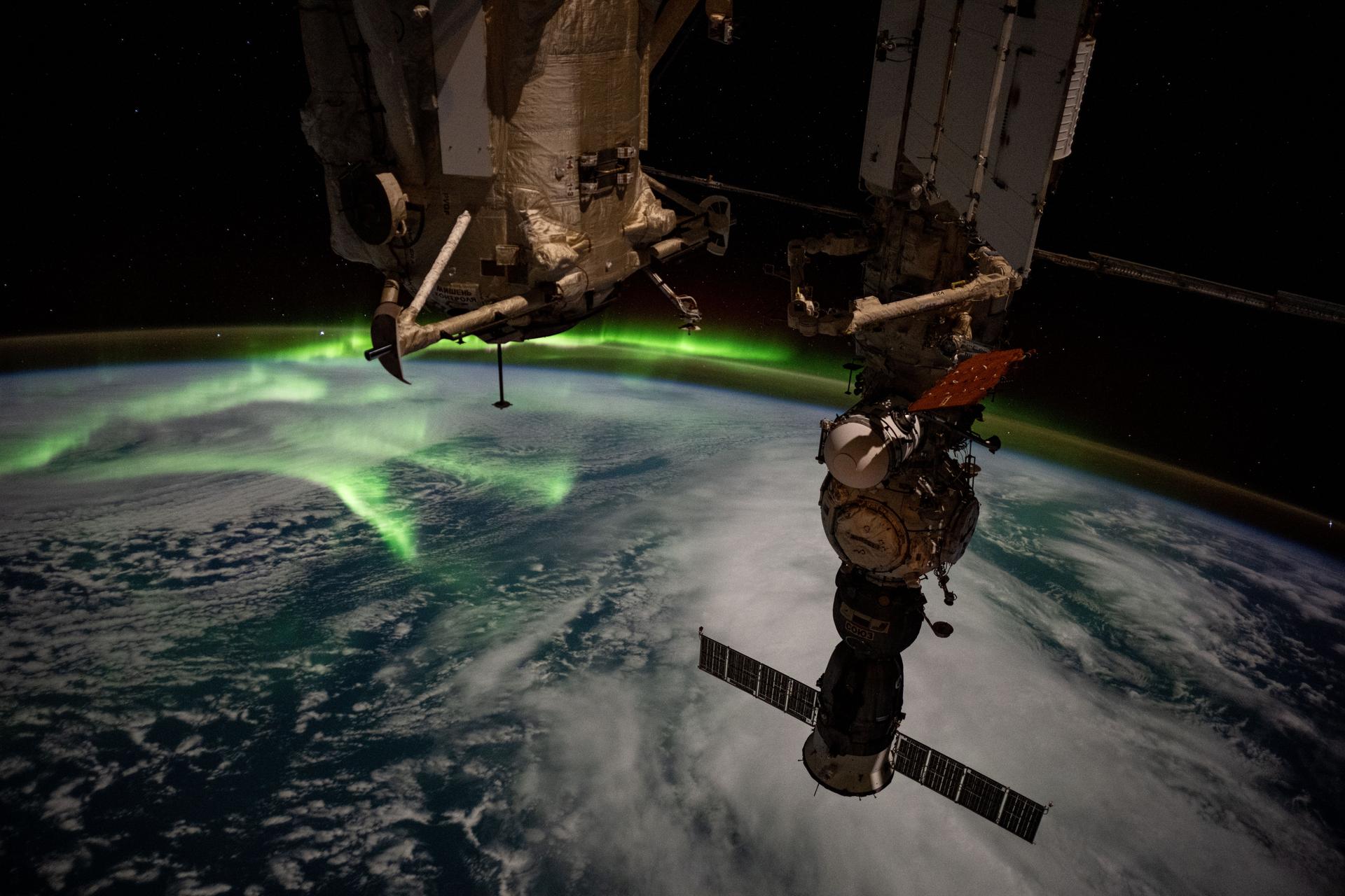 The aurora australis arcs back and forth above a partly cloudy Indian Ocean in this photograph from the International Space Station as it orbited 270 miles above in between Australia and Antarctica. At center top, is the Rassvet module, at lower right, is the Soyuz MS-27 crew ship docked to the Prichal module.
