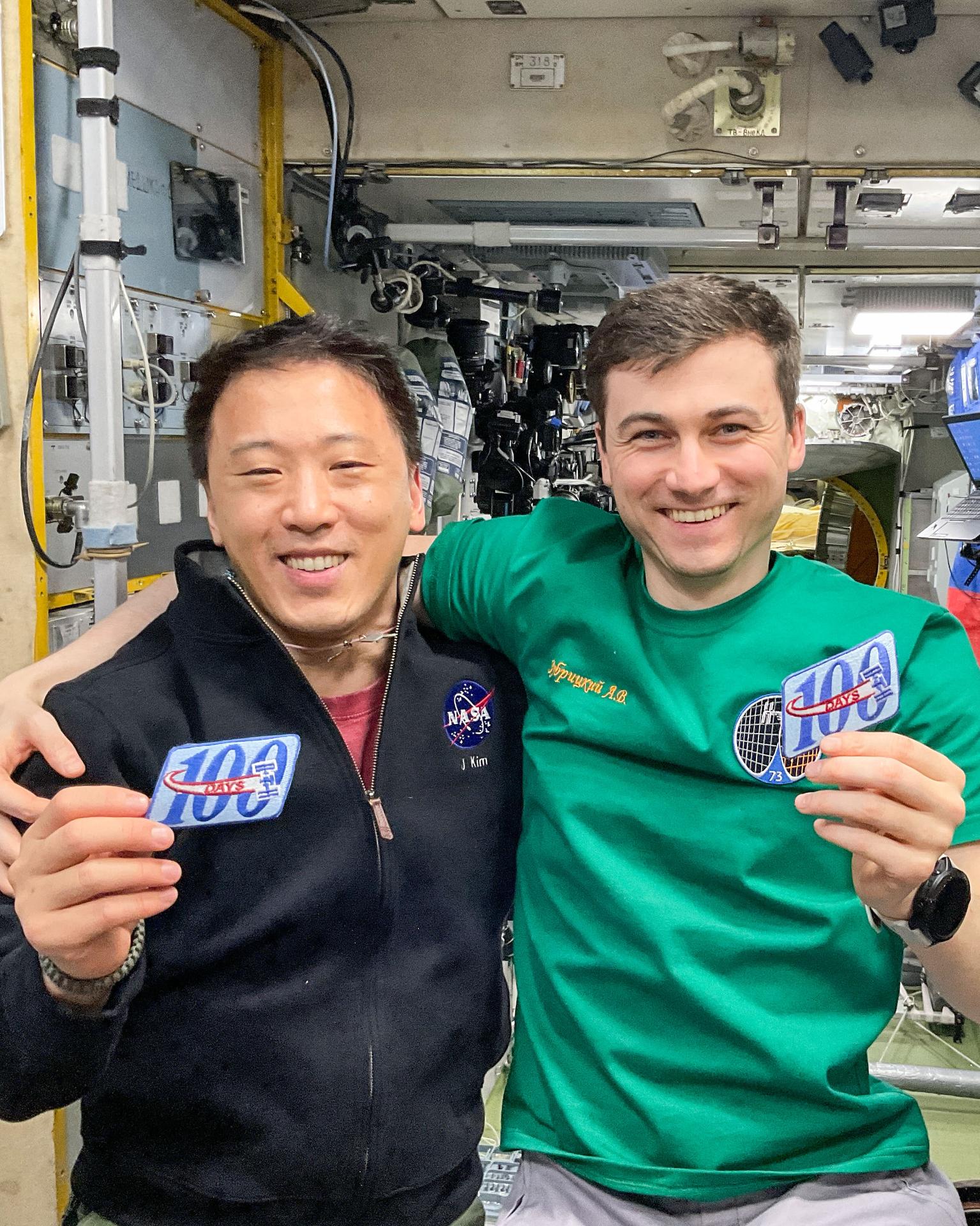 Expedition 73 Flight Engineers (from left) Jonny Kim of NASA and Alexey Zubritsky of Roscosmos, both first time space-flyers, show off patches marking a benchmark 100 days in space since their arrival aboard the Soyuz MS-27 spacecraft on March 8, 2025. The duo arrived aboard the Soyuz MS-27 with veteran Roscosmos cosmonaut Sergey Ryzhikov (not pictured) who is on his third mission aboard the orbital outpost.