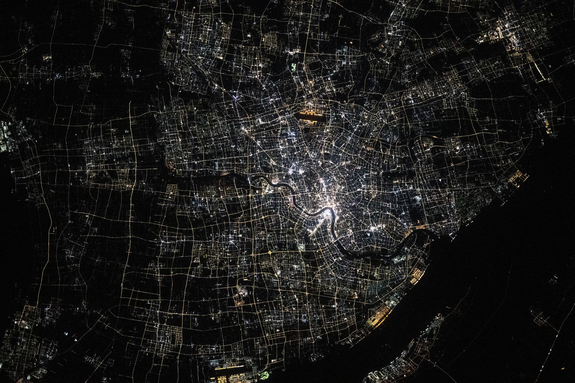 Shanghai, China—the nation's largest city with a metropolitan population of approximately 24.9 million—is seen from the International Space Station at around 9:55 p.m. local time. Located where the Yangtze River meets the Yellow Sea, the city glows with urban energy. Notable landmarks include Shanghai Hongqiao International Airport near the top center and Shanghai Pudong International Airport at the bottom. The Huangpu River winds through the heart of downtown, dividing the historic Bund from the futuristic skyline of Lujiazui.