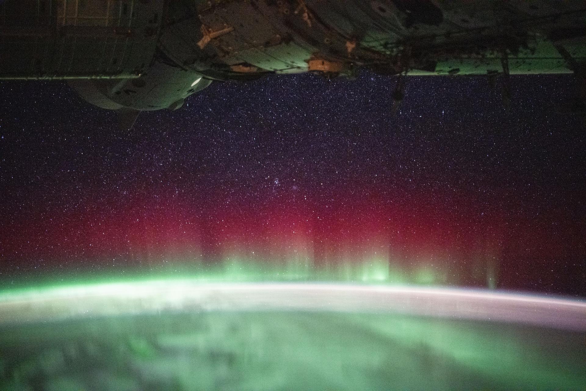 A vivid red and green aurora crowns Earth’s horizon over the southern Indian Ocean in this photo from the International Space Station as it orbited 270 miles above. At top left, a lit window reveals the docked SpaceX Dragon crew spacecraft docked to the orbital outpost's forward port on the Harmony module, set against a starry sky captured with long-exposure, low-light settings.