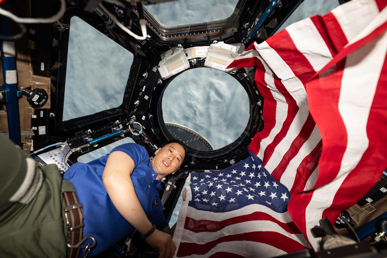 iss073e0420732 (July 4, 2025) --- NASA astronaut and Expedition 73 Flight Engineer Jonnny Kim poses for a portrait with the American flag on the Fourth of July, or Independence Day, inside the International Space Station's "window to the world," the cupola. The orbital outpost was soaring 268 miles above a cloudy Atlantic Ocean at the time of this photograph.