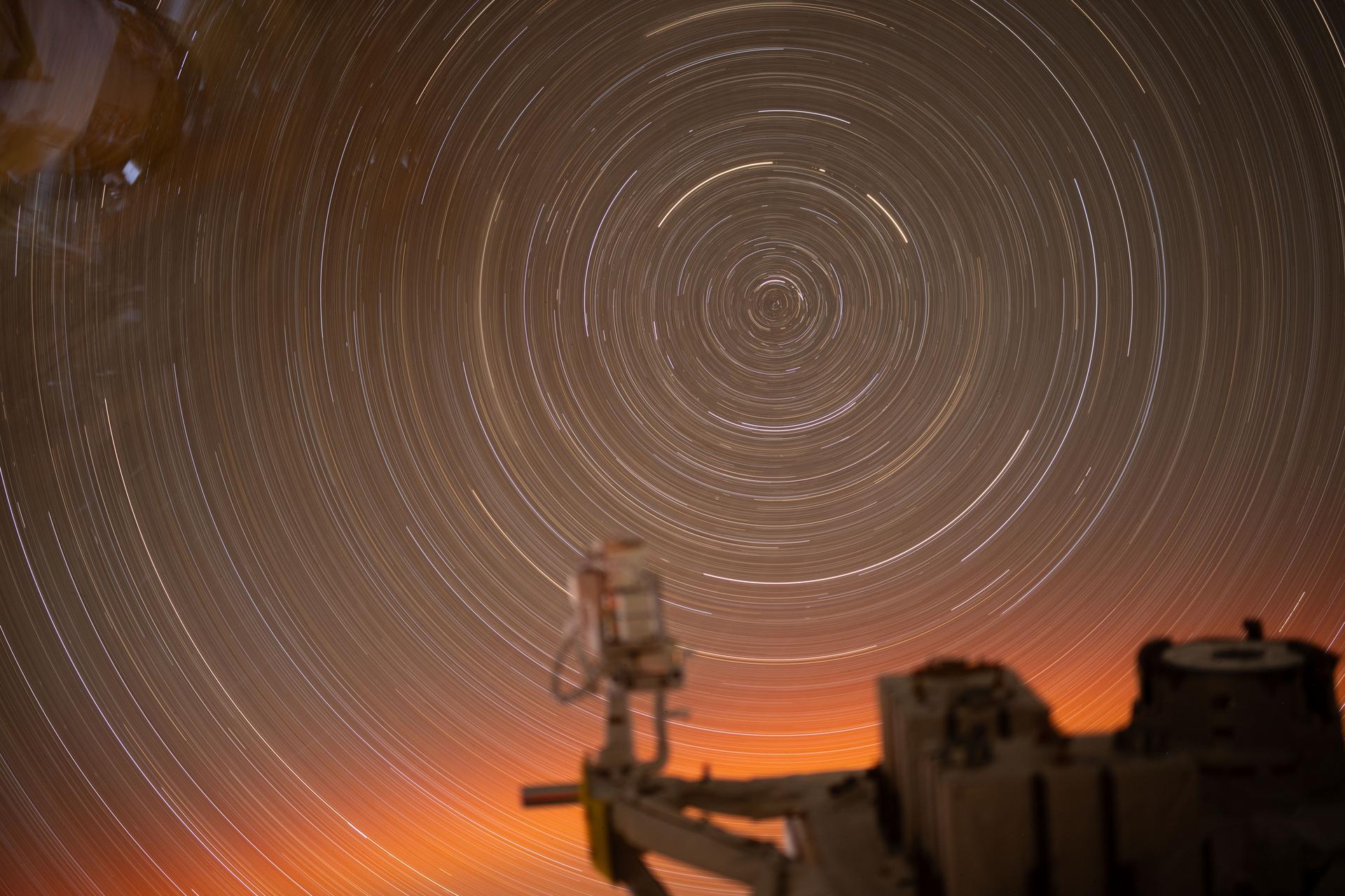 This long-exposure photograph, taken over 31 minutes from a window inside the International Space Station’s Kibo laboratory module, captures the circular arcs of star trails. In the foreground is a portion of Kibo’s Exposed Facility, where various payloads and experiments are mounted to be exposed directly to the vacuum of space.