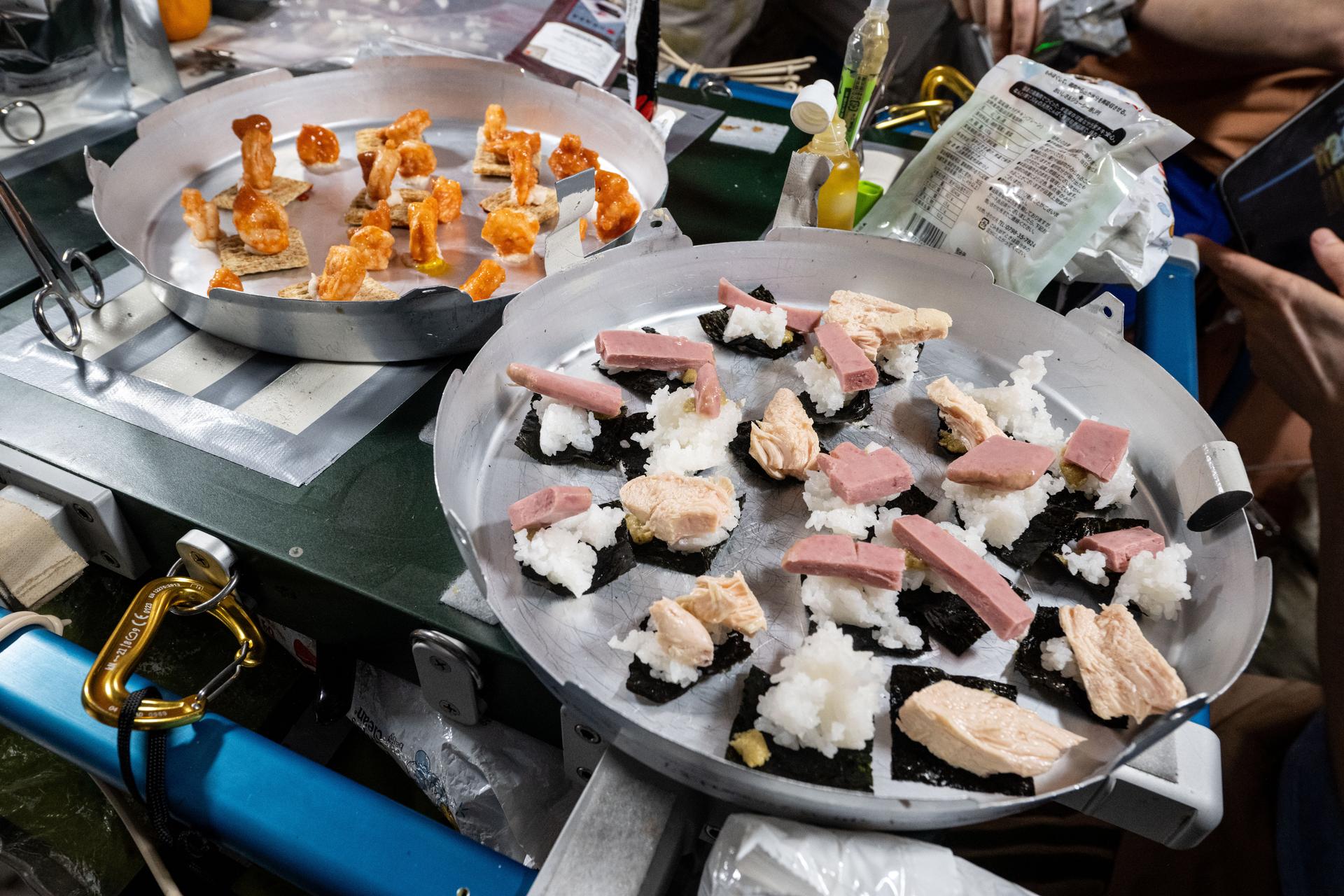 Dinner is served aboard the International Space Station! One tray features shrimp cocktail on whole grain wheat crackers, while the other holds sushi made with seaweed, spam, tuna, and rice. Both trays are secured with Velcro strips to keep them stable inside the Unity module’s galley. The shrimp and crackers are held in place by condiments, while the sushi stays put thanks to surface tension from its moisture.