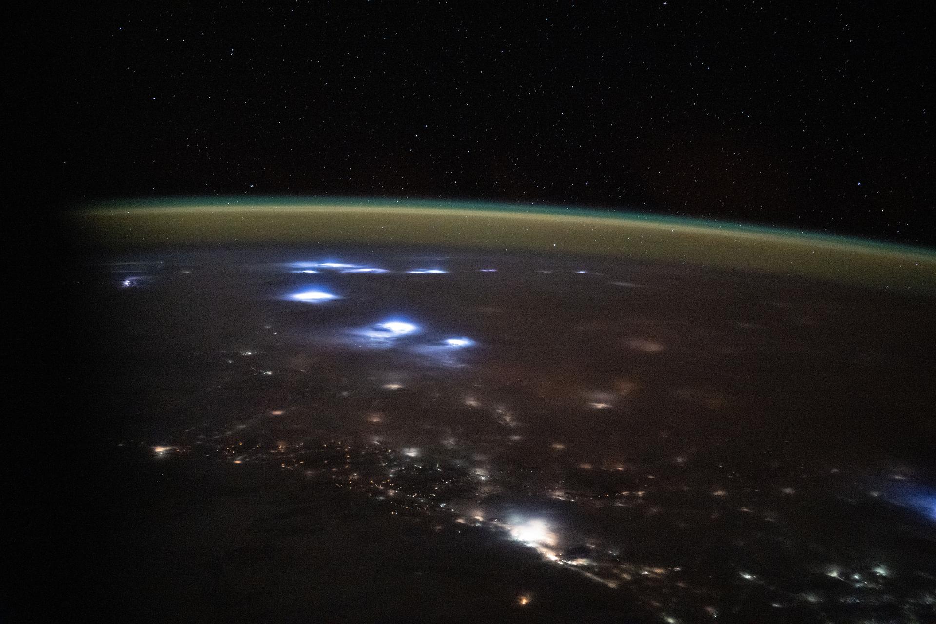 Warm, diffuse city lights and bright lightning storms illuminate the cloudy nighttime landscape of western Africa in this image captured from the International Space Station. A soft yellow-green atmospheric glow, created by atoms and molecules releasing energy absorbed from solar radiation earlier in the day, blankets Earth’s horizon. The orbital outpost was soaring 260 miles above the Atlantic Ocean at approximately 11:05 p.m. local time when this photograph was taken.