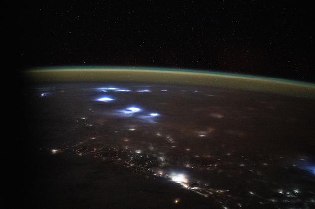 NASA image: Warm, diffuse city lights and bright lightning storms illuminate the cloudy landscape