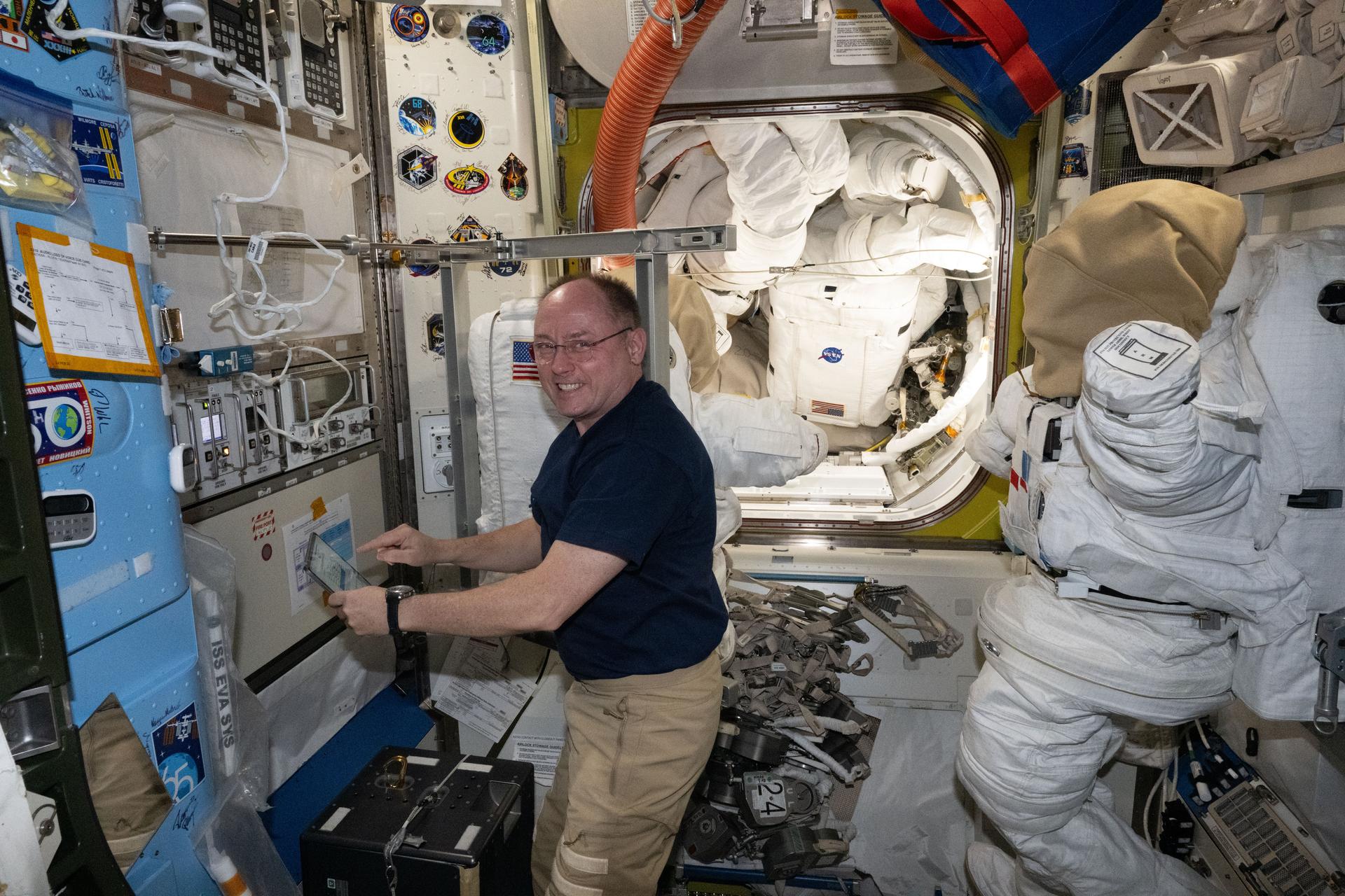 NASA astronaut and Expedition 73 Flight Engineer Mike Fincke performs maintenance tasks inside the International Space Station’s Quest airlock. Behind him, a U.S. spacesuit is secured to a metallic donning structure on the airlock wall, with its gloves removed and a protective cover placed over the helmet.