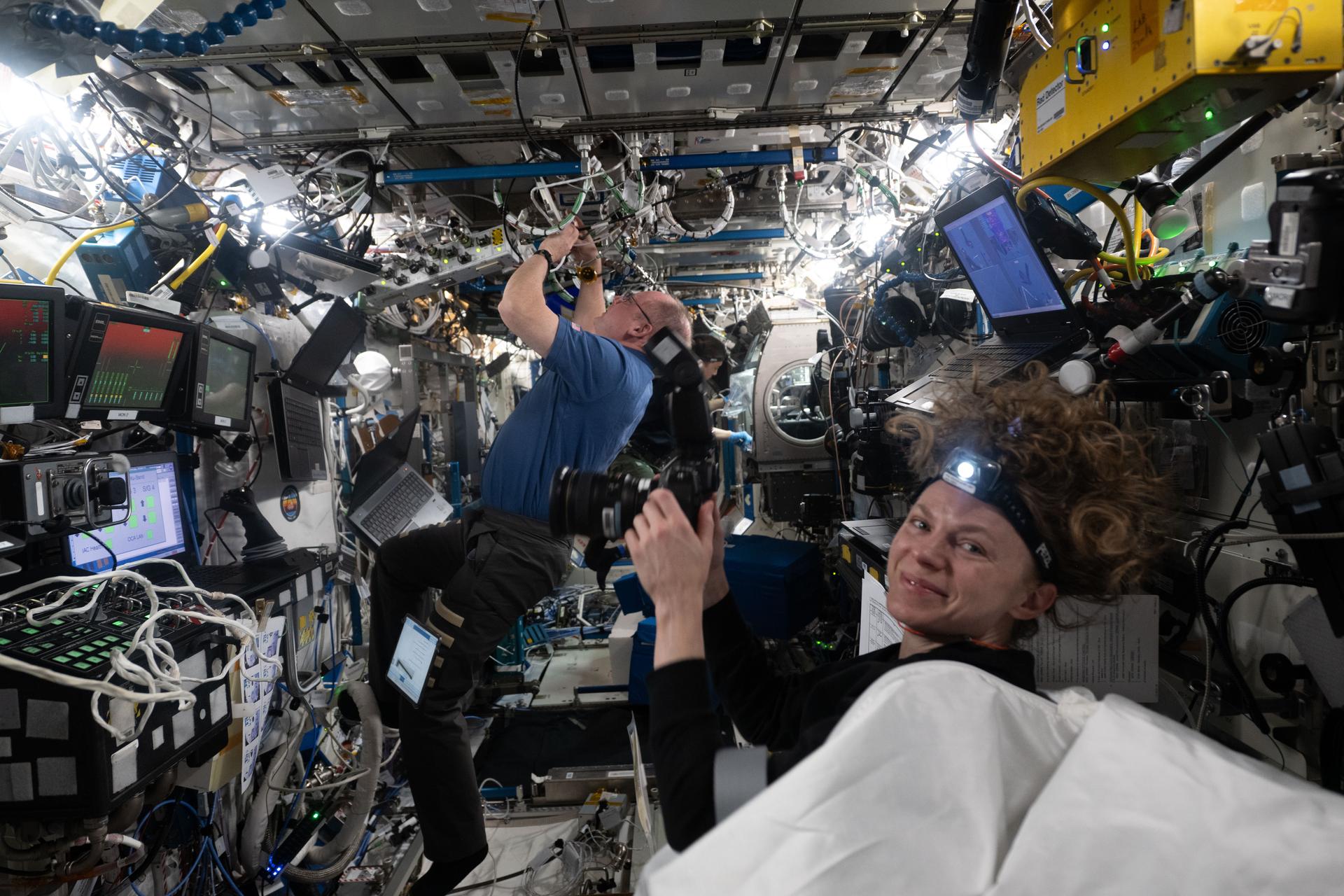 NASA astronaut Zena Cardman prepares to photograph NASA astronauts Mike Fincke and Jonny Kim during scientific and maintenance operations inside the International Space Station's Destiny laboratory module.