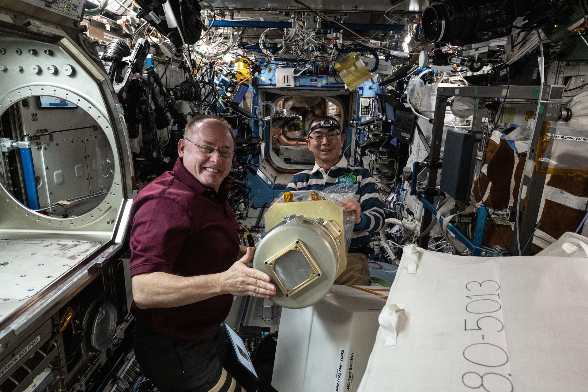 Fincke wears a maroon shirt and smiles as he holds a metallic cylindrical tank with a square outcrop on its flat end. Yui, wearing a striped white and blue shirt, stands next to Fincke and removes a bag from the tank. The surrounding walls of the space station have many cables and electronics embedded into the walls.