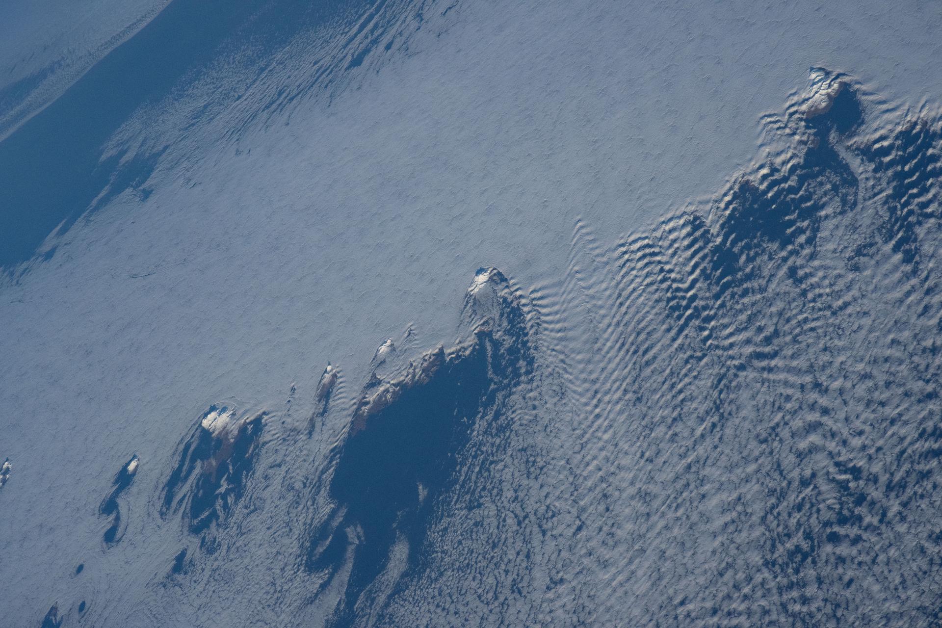 Snow-capped mountain tops of Alaska's Aleutian Islands pierce through the clouds, disturbing the air and creating wave-like patterns known as altocumulus clouds. This photograph was taken from the International Space Station as it orbited 264 miles above the North Pacific Ocean.