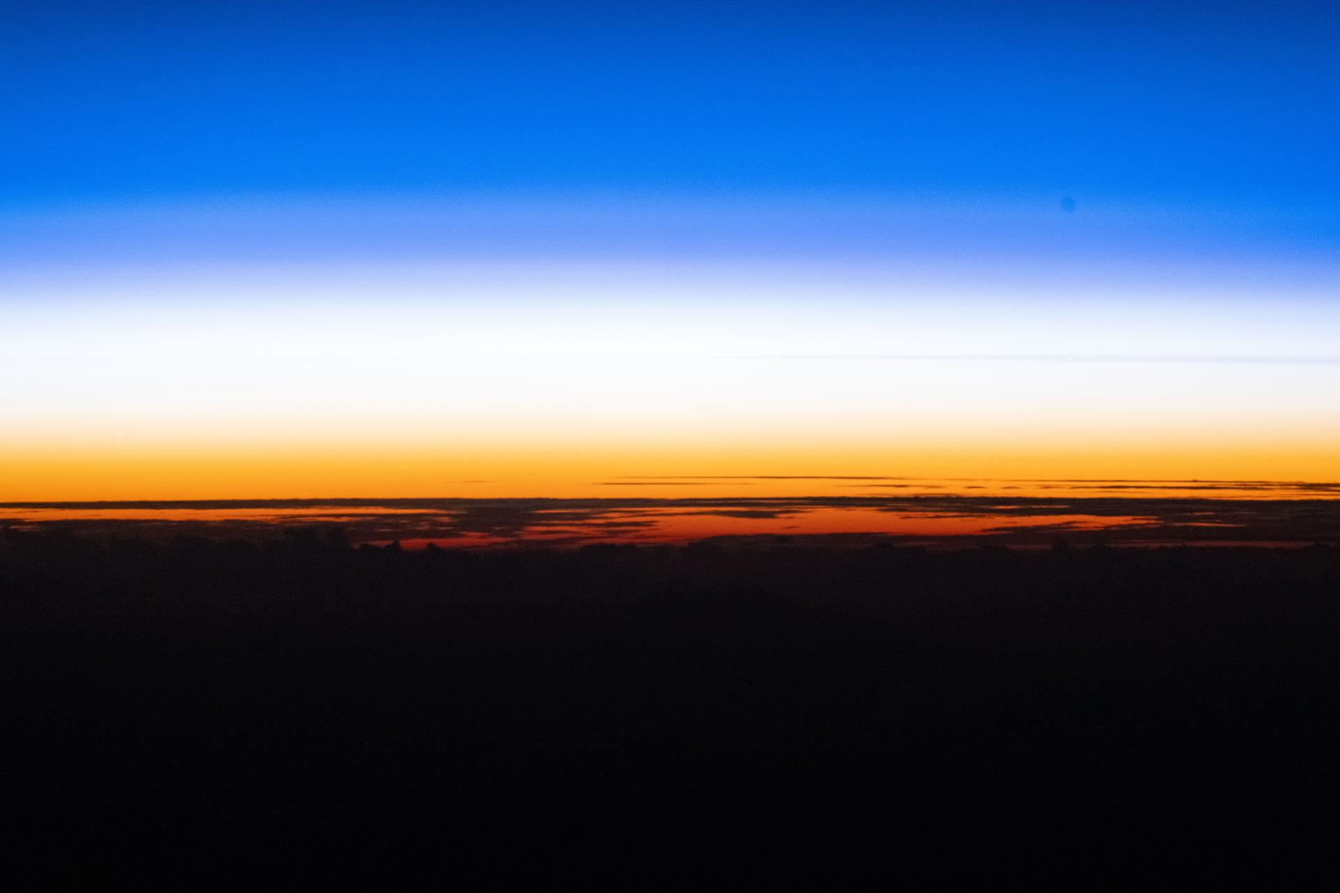 An orbital sunset silhouettes the cloud tops above the Pacific Ocean as the atmosphere along the horizon fades from orange to white, then to blue, in this photograph taken from the International Space Station while it orbited 262 miles above Earth. Credit: NASA/Chris Williams