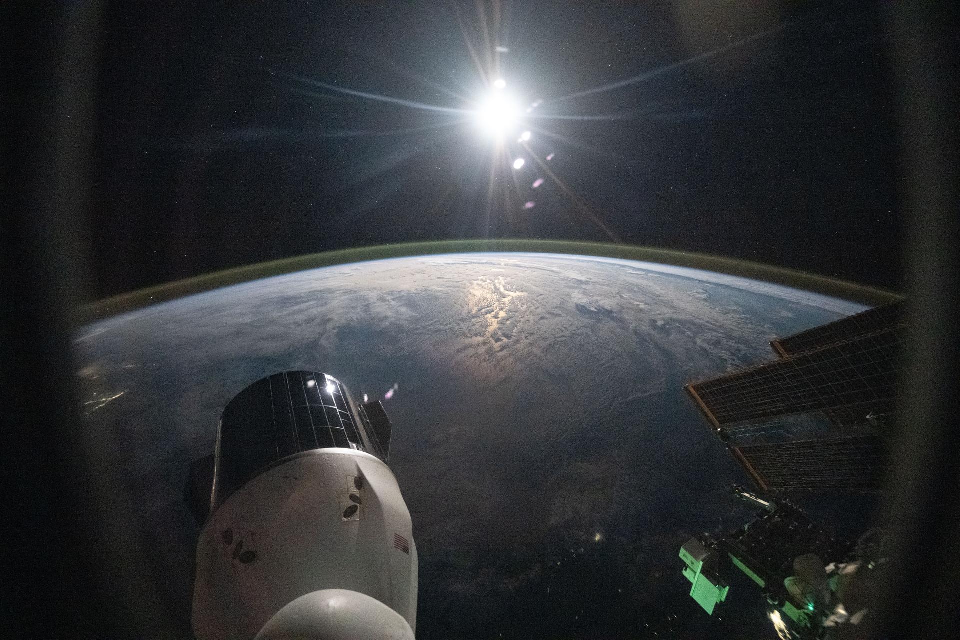The sun begins setting above Earth's atmospheric glow blanketing a cloudy Atlantic Ocean. The International Space Station was orbiting 260 miles above the Earth off the coast of Florida when this photograph was taken. In the foreground, are a SpaceX Dragon spacecraft (lower left) and a set of the orbital outpost's solar arrays (right).