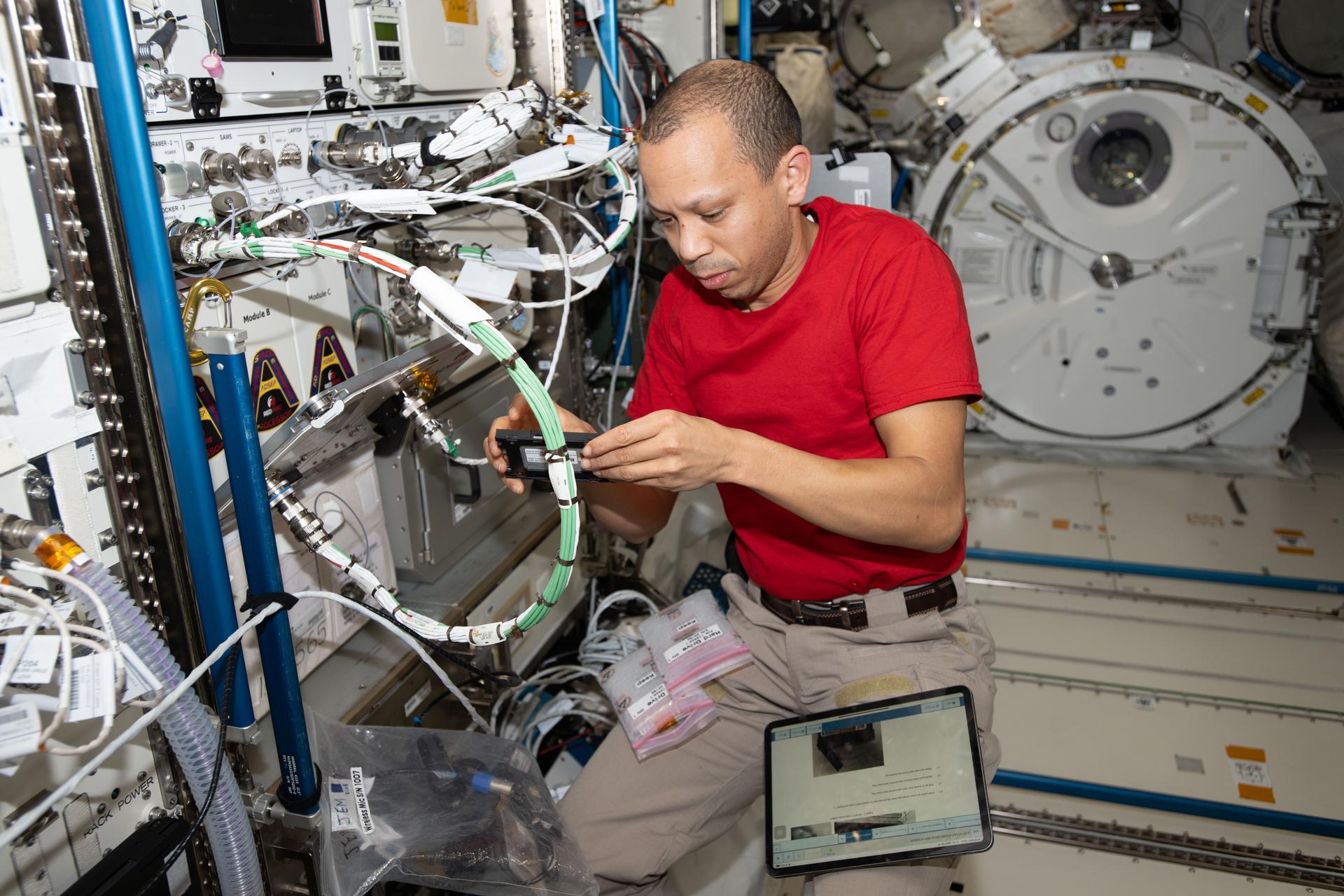 NASA astronaut Chris Williams holds a small, rectangular hard drive as he works to swap computer components for a protein crystal growth investigation. A tablet is secured to one of his legs, while two plastic bags are attached to the other. Bundles of cables float next to him and a circular hatch is visible in the background.