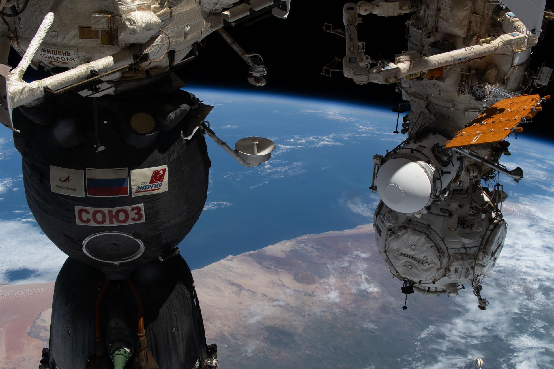 iss074e0325203 (Feb. 24, 2026) --- Namibia's Brandberg Massif, near the center of the photograph, appears as a nearly perfect circular formation just inland from the Atlantic coast, and easy to mistake for a meteorite impact crater when seen from space. At left, is the Soyuz MS-28 crew spacecraft docked to the Rassvet module. At right, the Prichal docking module is attached to the Nauka science module. The International Space Station was orbiting 266 miles above southern Africa at the time of this photograph. Credit: NASA/Jessica Meir