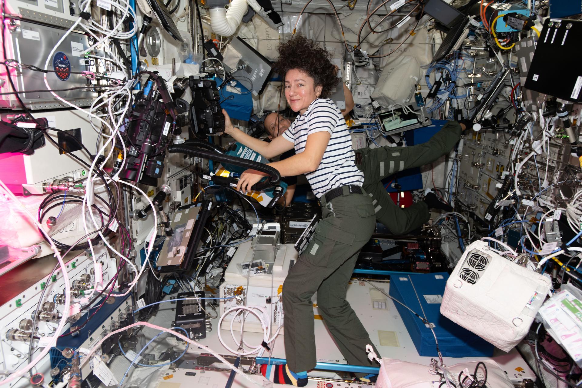 iss074e0503707 (April 22, 2026) --- NASA astronauts Jessica Meir and Chris Williams (in the background) work on separate tasks inside the International Space Station’s Columbus laboratory module. Meir swapped scientific hardware inside the BioLab research incubator to study how DNA damaged by space radiation repairs itself. Williams installed new laptop software and checked computer connections to the station’s Wi‑Fi system. Credit: NASA/Jack Hathaway