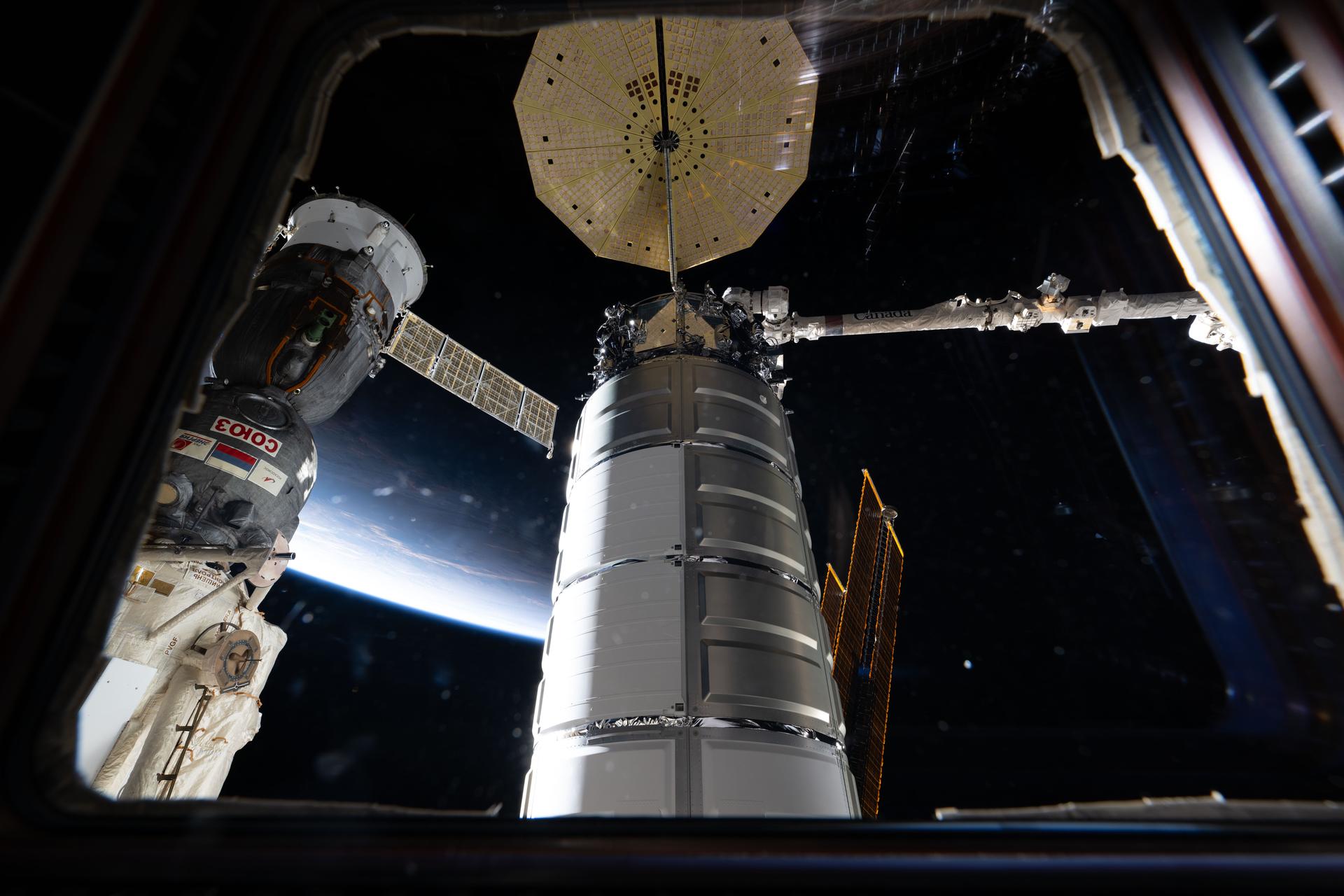 Two spacecraft attached to the International Space Station are seen through a window in the cupola. In the foreground, is Northrop Grumman's Cygnus XL cargo spacecraft, in the grips of the Canadarm2 robotic arm, after its capture and installation to the Unity module. At left, is the Soyuz MS-28 crew spacecraft docked to the Rassvet module.