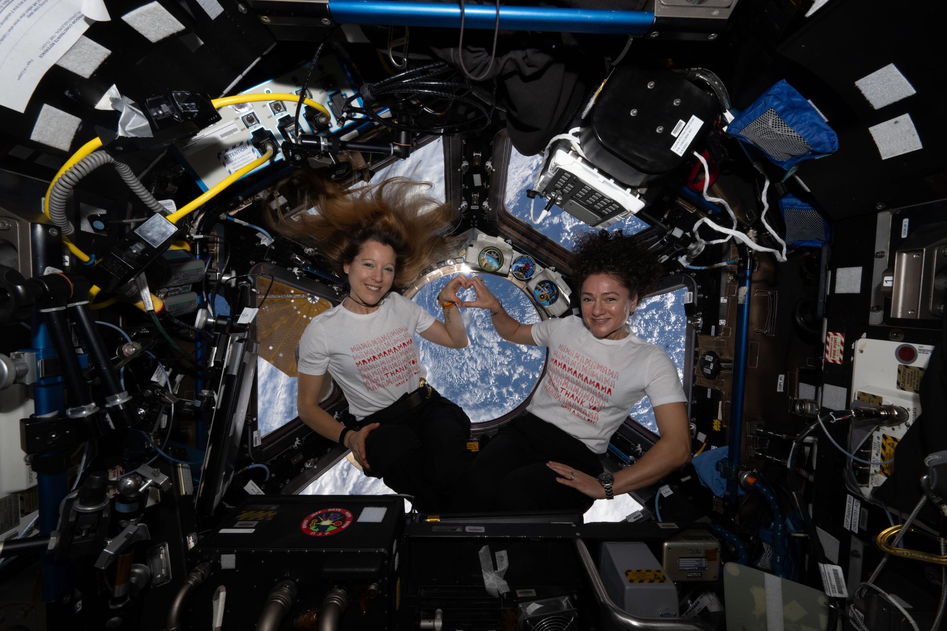 From left, Expedition 74 flight engineers Sophie Adenot of ESA (European Space Agency) and Jessica Meir of NASA pose for a portrait inside the International Space Station’s cupola on Mother’s Day. Adenot, who has a son, and Meir, who has a daughter, join hands to form a heart and wear T‑shirts celebrating motherhood.