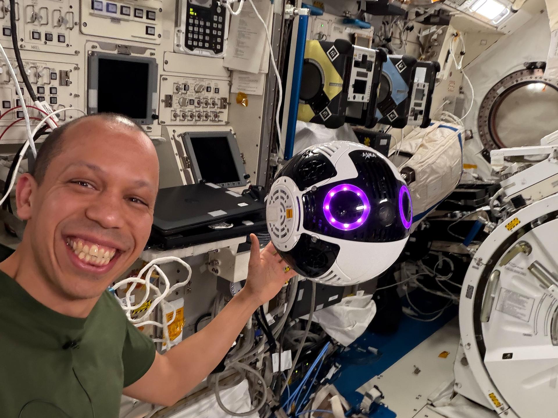 NASA astronaut and Expedition 74 flight engineer Chris Williams poses aboard the International Space Station’s Kibo laboratory module with a free‑flying robotic camera developed by JAXA (Japan Aerospace Exploration Agency) that could assist astronauts with video and photography activities.