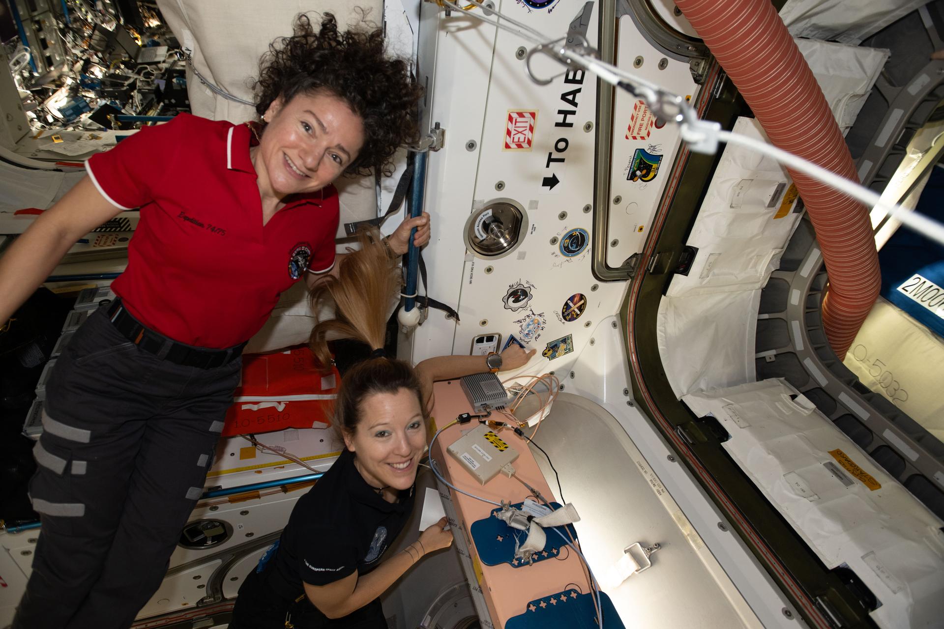 Expedition 74 flight engineers, from left, Jessica Meir of NASA and Sophie Adenot of ESA (European Space Agency), show off a Cygnus mission sticker for Northrop Grumman's 24th cargo mission to the International Space Station. The sticker is affixed to the vestibule between the orbital outpost's Earth-facing port on the Unity module and the company’s Cygnus XL cargo spacecraft.