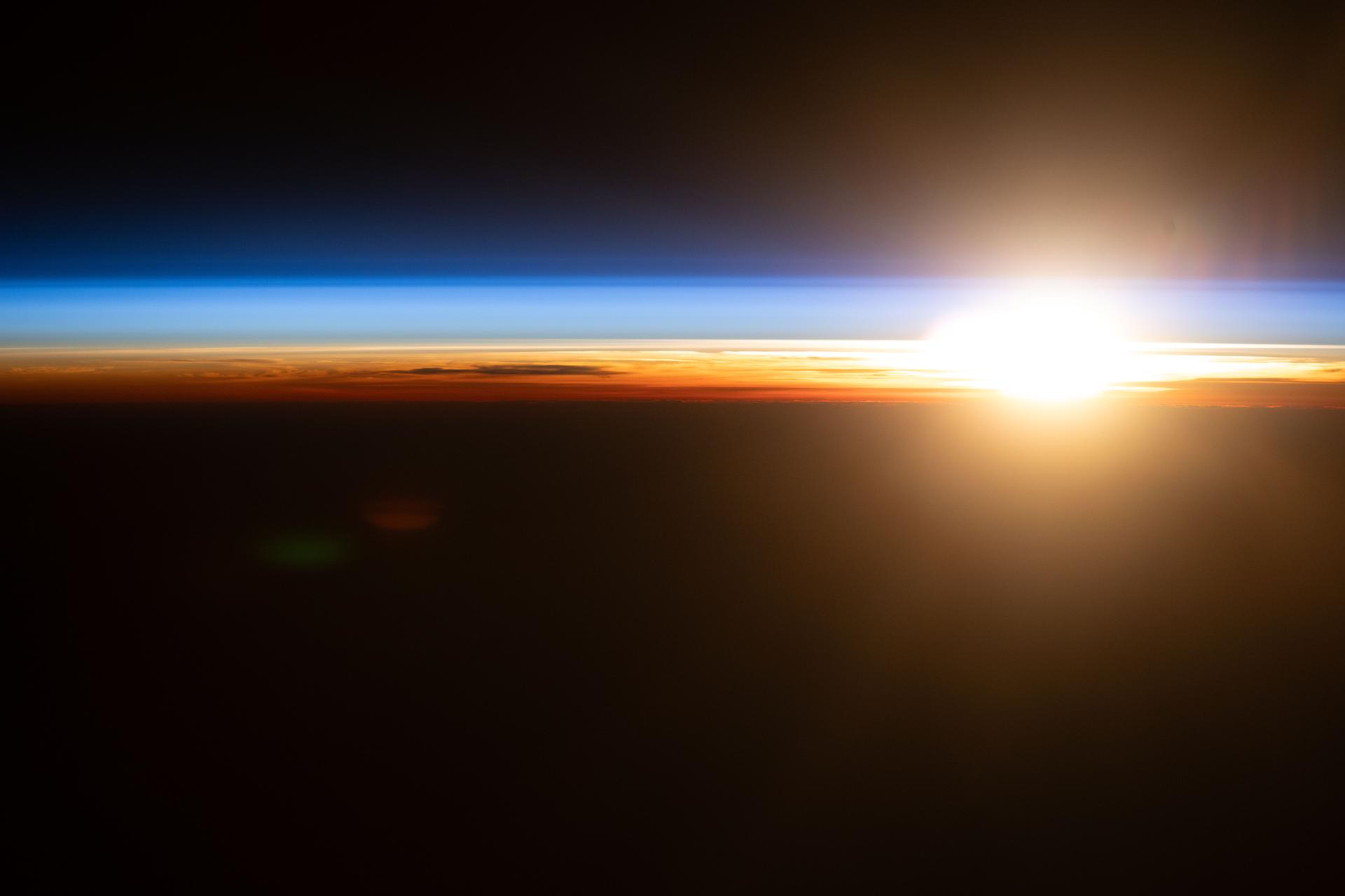 An orbital sunrise above the Pacific Ocean illuminates Earth's atmosphere, revealing cloud tops and orange and blue hues outlining the planet's horizon. The International Space Station was orbiting 263 miles above the Hawaiian island chain at approximately 4:09 a.m. local time when this photograph was taken.