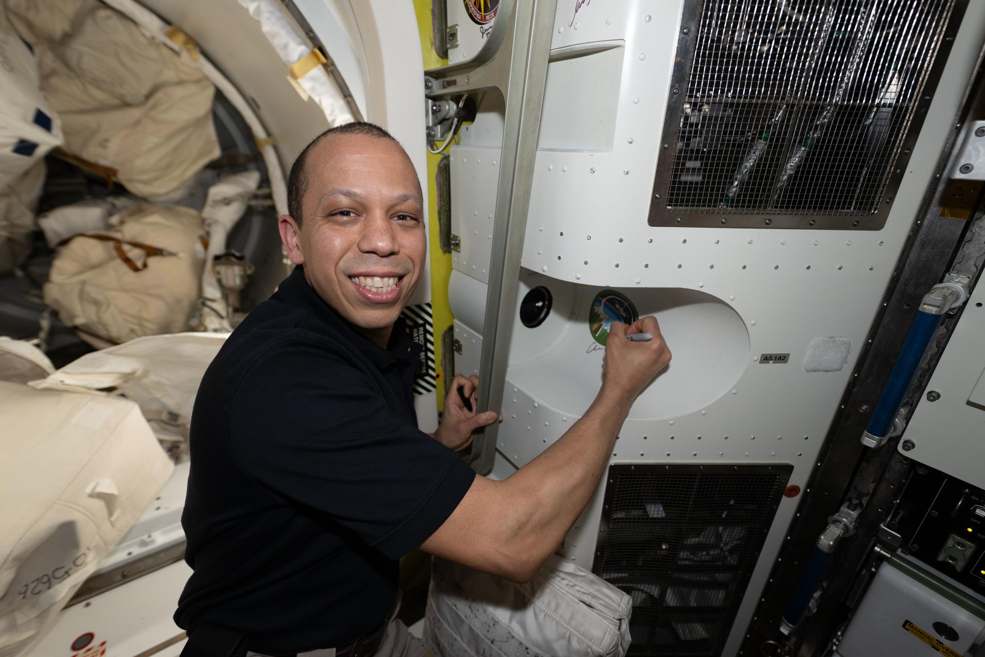NASA astronaut and Expedition 74 flight engineer Chris Williams poses for a portrait while signing his autograph on the wall around the Expedition 74 mission sticker inside the International Space Station’s Quest airlock.