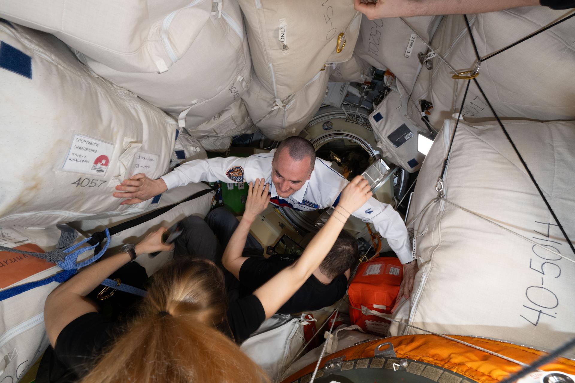 Roscosmos cosmonaut Pyotr Dubrov (top) enters the International Space Station shortly after docking to the Prichal module inside the Soyuz MS-29 spacecraft. Welcoming Dubrov are Expedition 74 flight engineers Sophie Adenot of ESA (European Space Agency) and Jack Hathaway of NASA.