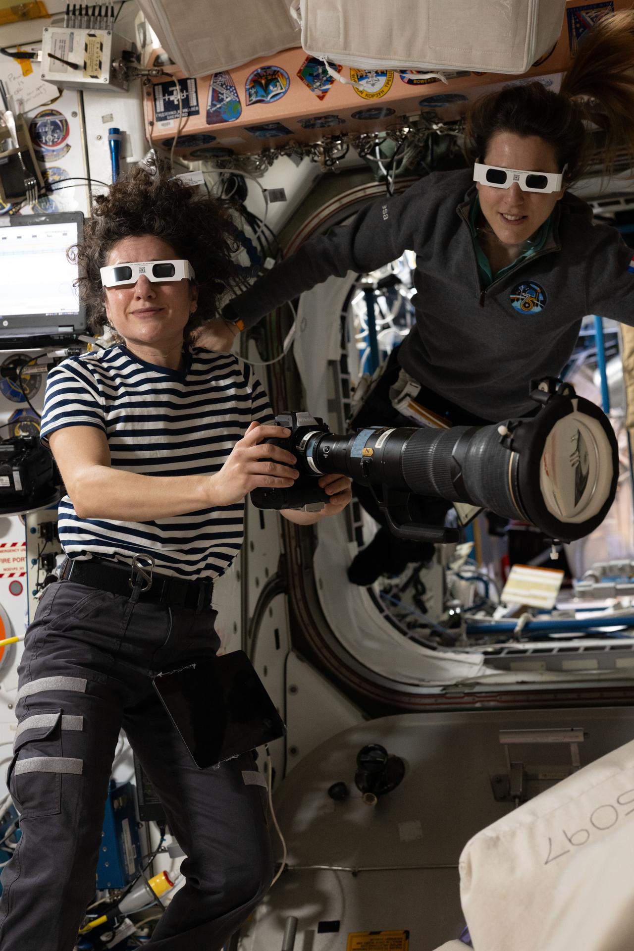 From left, Expedition 75 commander Jessica Meir of NASA and flight engineer Sophie Adenot of ESA (European Space Agency) wear solar eclipse glasses to protect their eyes while photographing a partial solar eclipse from the International Space Station. Meir is holding a Nikon Z9 camera with an 800mm lens, a 1.4x teleconverter, and a solar filter.