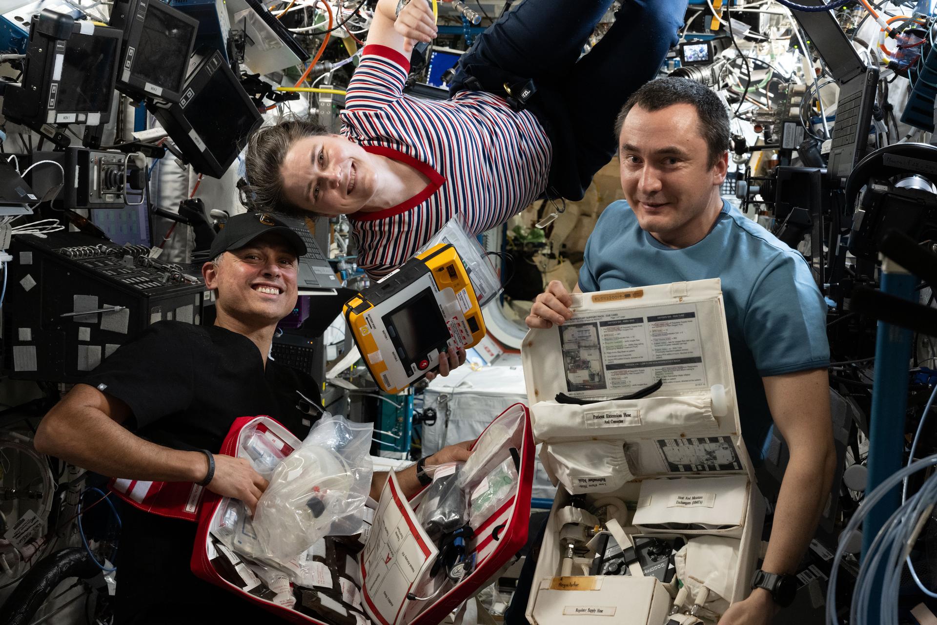 Expedition 75 flight engineers Anil Menon of NASA and Anna Kikina and Pyotr Dubrov of Roscosmos smile for a portrait as they check out emergency medical equipment inside the International Space Station's Destiny laboratory module.