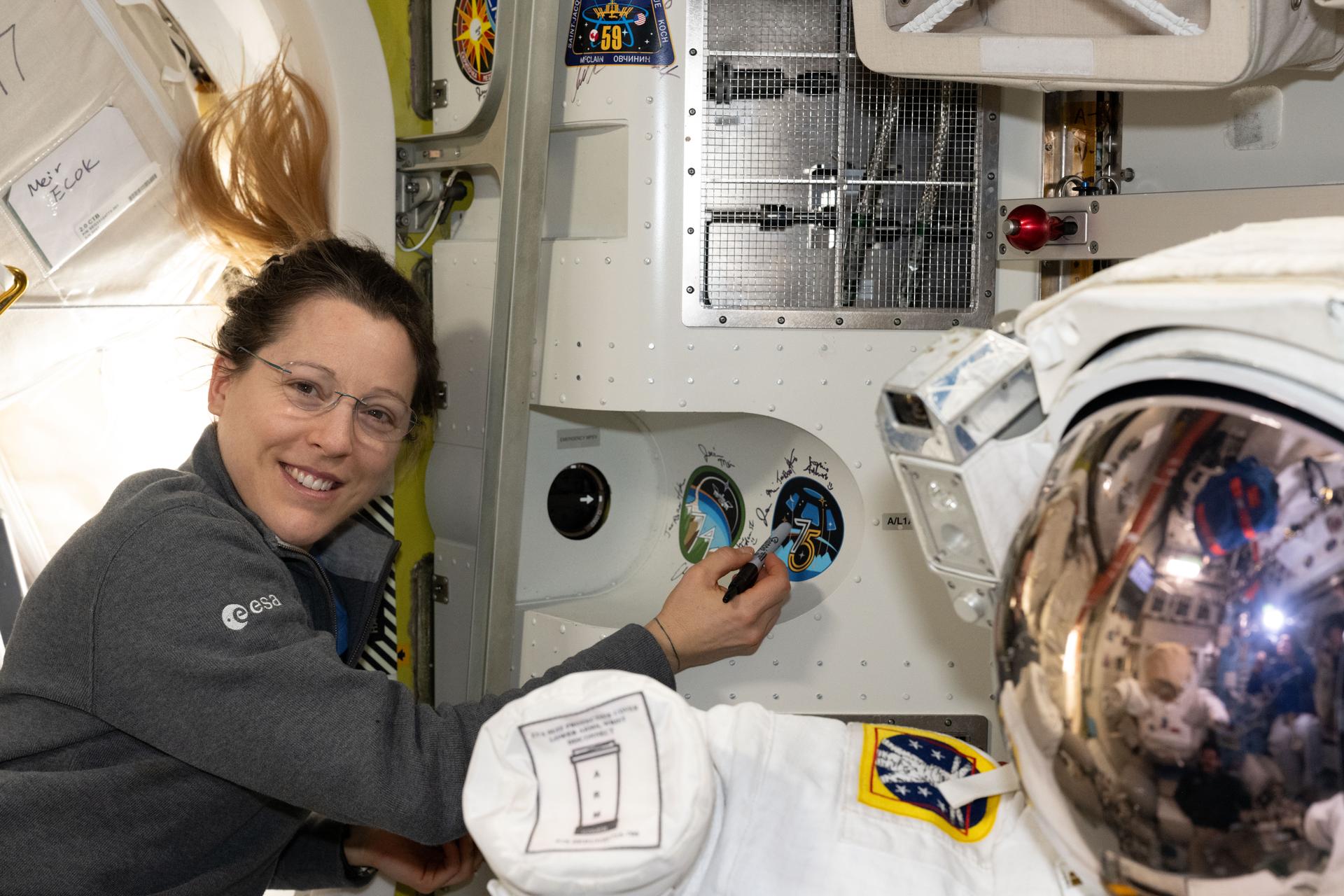 iss075e0144645 (Sept. 23, 2026) --- ESA (European Space Agency) astronaut and Expedition 75 flight engineer Sophie Adenot points at her mission's sticker affixed to the inside of the International Space Station's Quest airlock where spacesuits are staged and prepared for spacewalks. Credit: NASA/Anil Menon