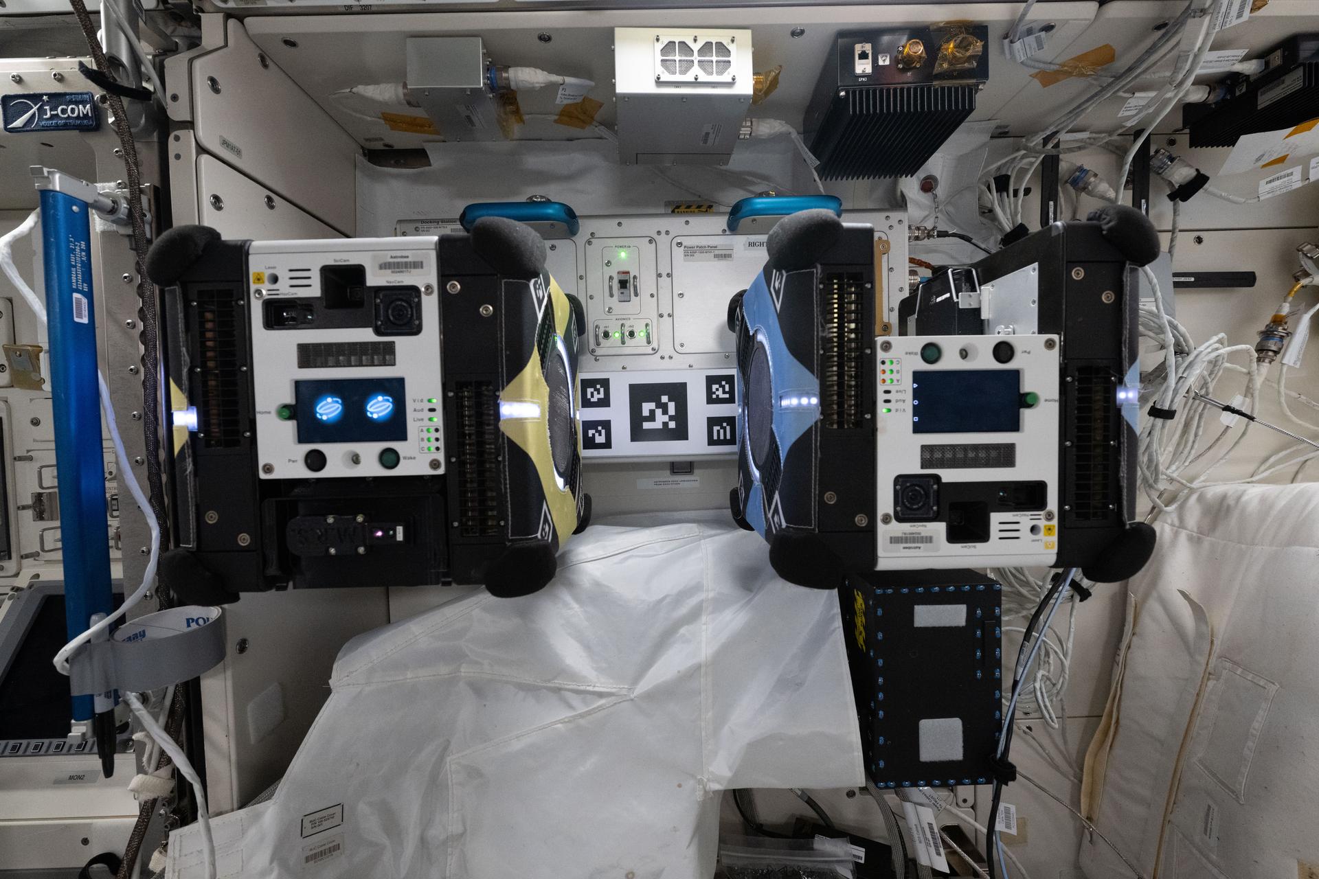 A pair of toaster-sized Astrobee free-flying robotic assistants are pictured attached to a docking station inside the International Space Station's Kibo laboratory module. The Astrobee docking station provides recharging power, cooling airflow, and data connectivity for the robotic free-flyers.