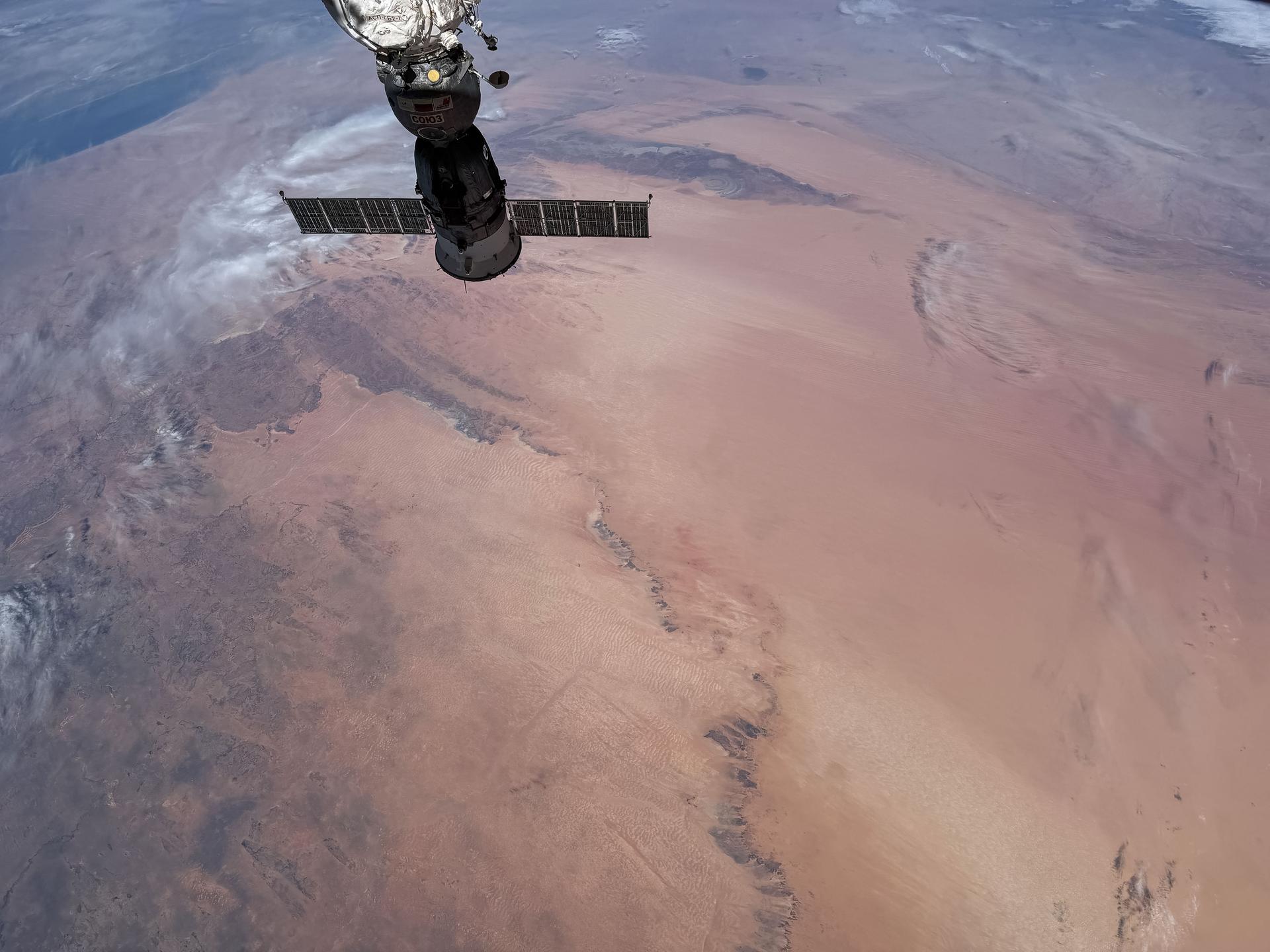The Soyuz MS‑29 crew spacecraft is pictured docked to the Prichal module as the International Space Station orbits above Africa. At the top right of the Soyuz is the Richat Structure, also known as the Eye of the Sahara, a geological formation located in Mauritania. The Richat Structure formed when underground rock was pushed upward into a dome, and erosion later revealed its layers as giant circular rings.
