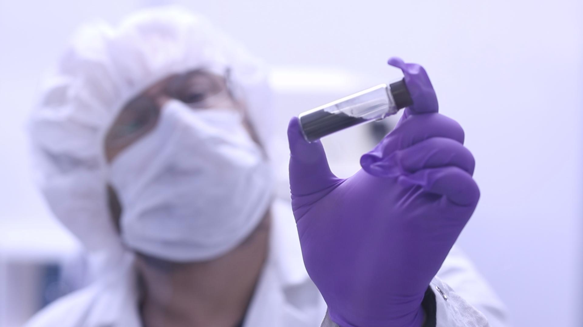 A man wearing a white lab coat, purple gloves, and a white hair covering, holds a vial containing a small sample of asteroid Bennu in front of him. The sample is gray. The depth of field is shallow, so his hand and the vial are in focus, and the rest of him is slightly out of focus.