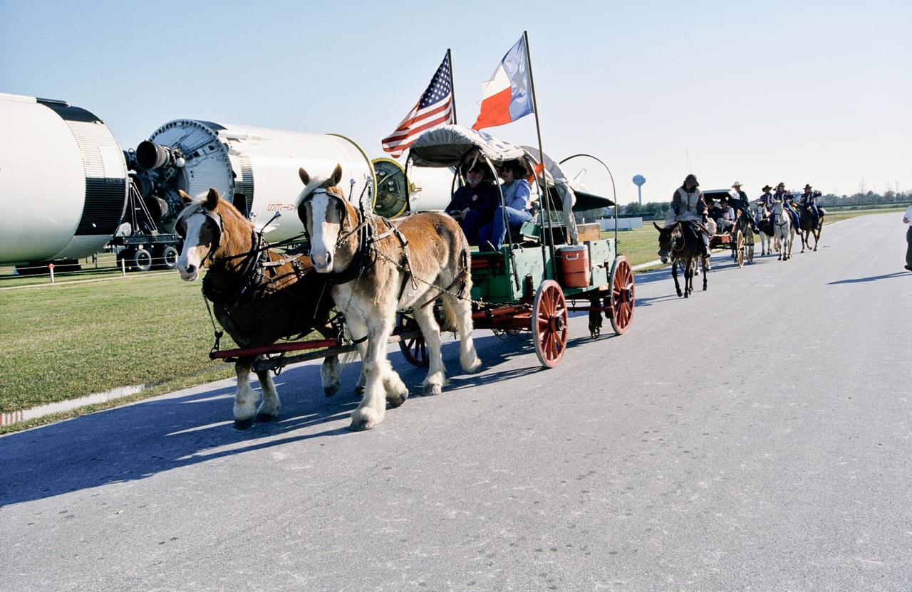 Photographic documentation showing a Cowboy Parade at JSC, part of Rodeo Houston events. Photo taken at Rocket Park with Saturn V rocket exhibit in the background.