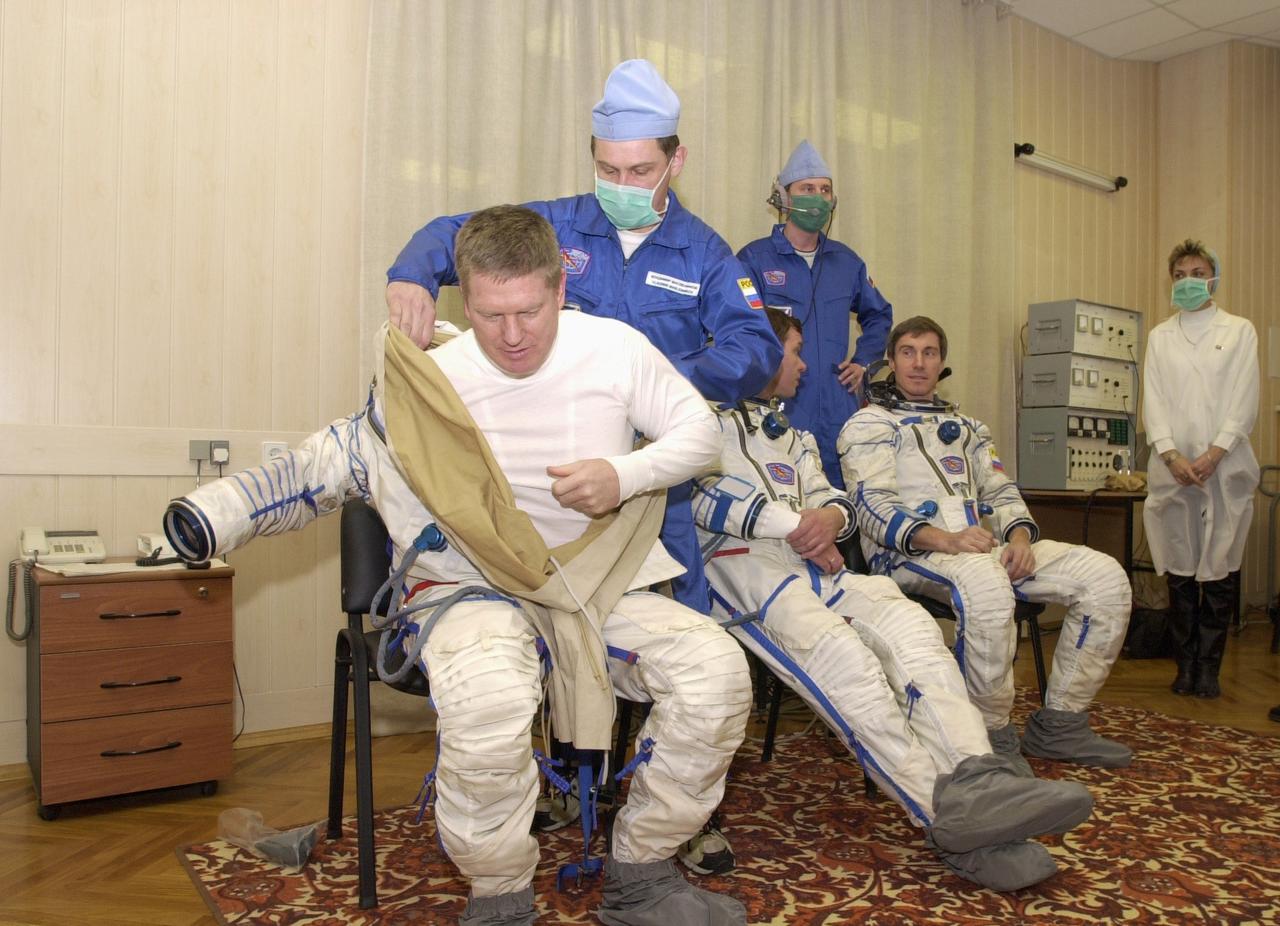 JSC2000-E-27081 (20 October 2000) --- Expedition One commander William M. (Bill) Shepherd gets help with his Sokol suit prior to a simulation at Baikonur. Also  donning suits are Soyuz pilot Yuri P. Gidzenko (seated at center) and flight engineer Sergei K. Krikalev.