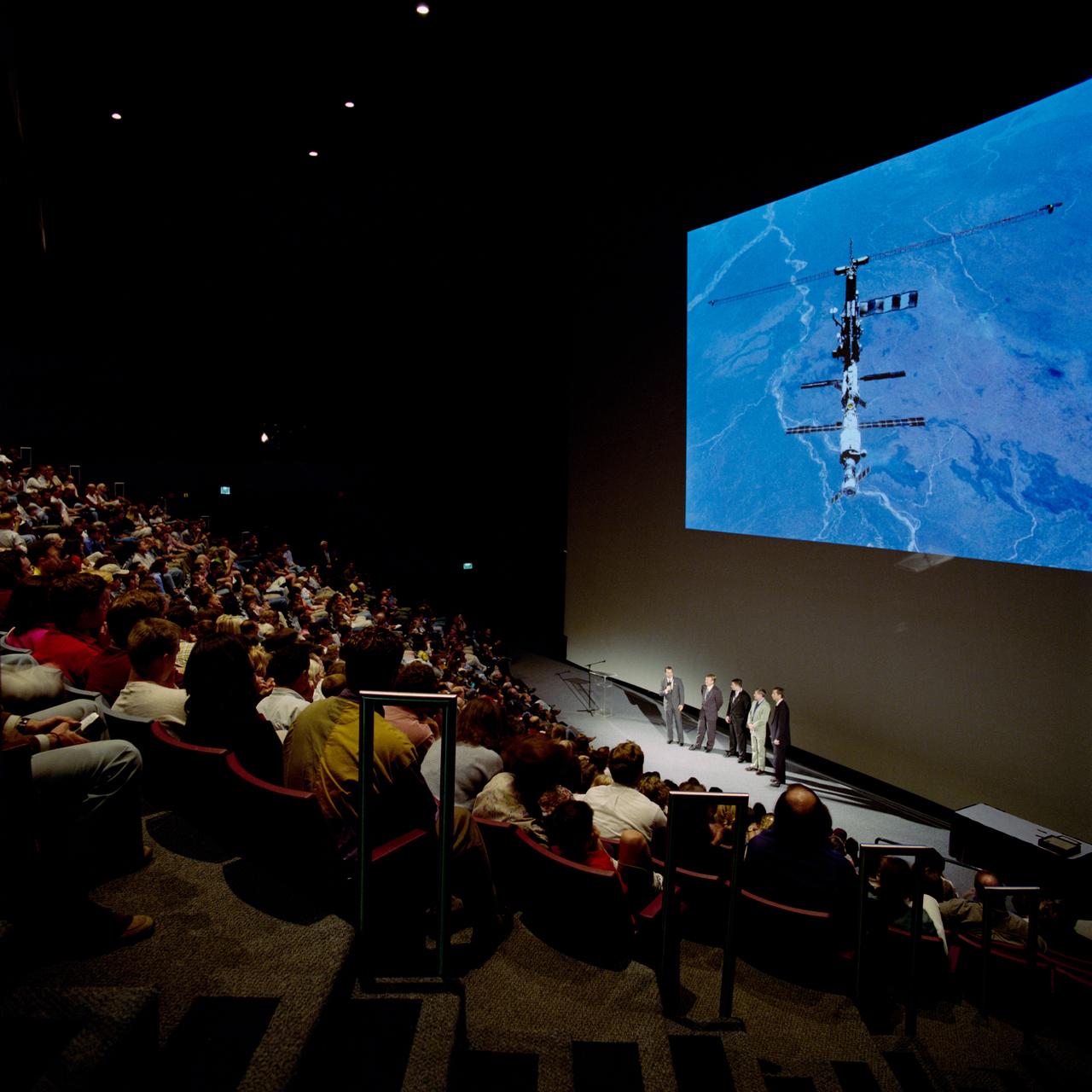 View of individuals in audience watching the International Space Station (ISS) on large screen.