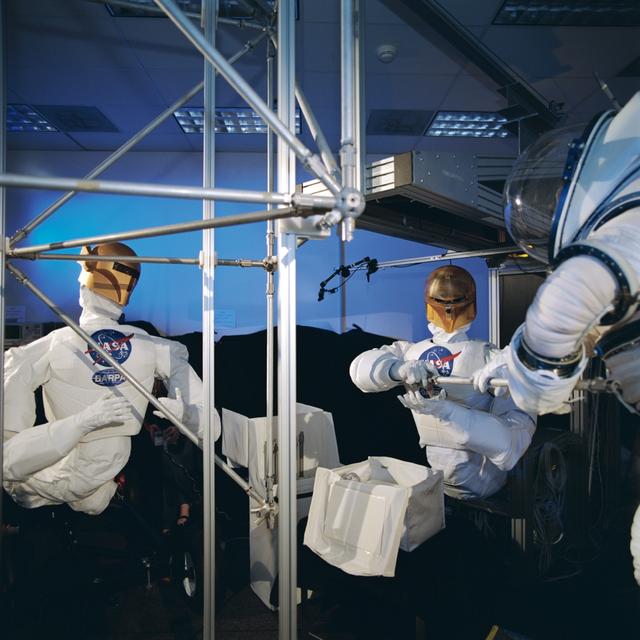 JSC2003-00300 (June 2003) --- Two Robonauts work hand-in-hand with astronaut Nancy J. Currie, partially visible at right edge of frame, during an evaluation at the Johnson Space Center to determine the capabilities of human beings and robots to perform certain extravehicular tasks.